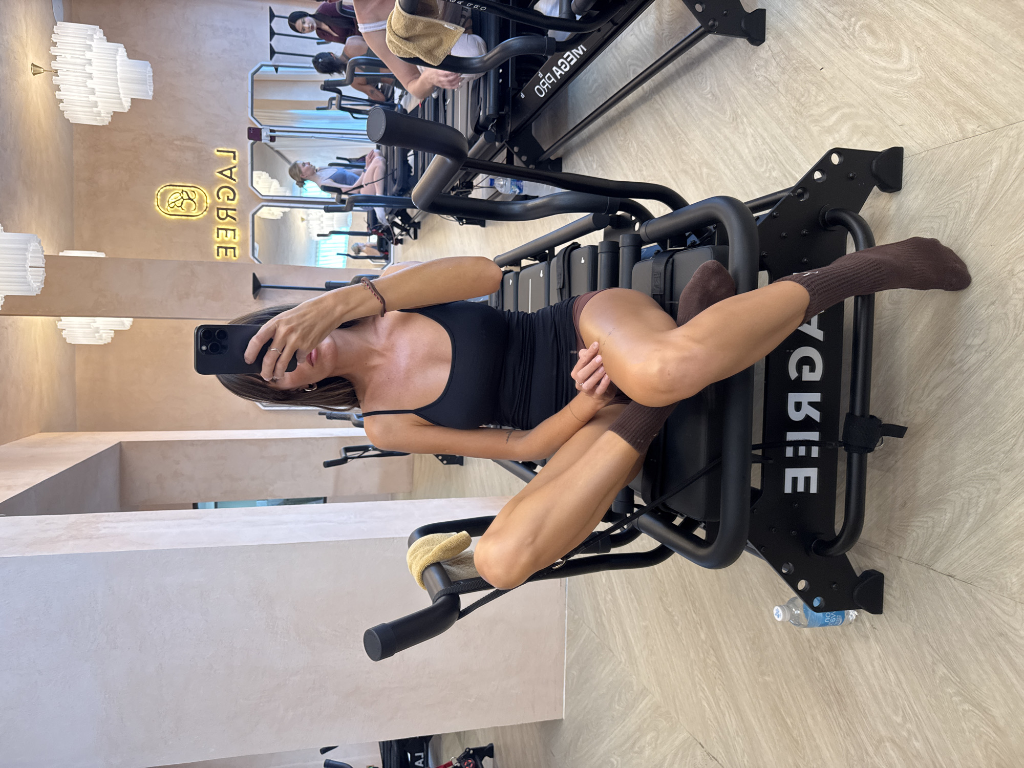 A woman is lying on a Lagree fitness machine inside what appears to be a fitness studio. She is wearing a black tank top, brown socks, and a bracelet. She is holding a black smartphone and taking a selfie. The Lagree machine is black, and there are other similar machines in the background. A partially visible sign with the word "LAGREE" is on the wall. There is a water bottle on the floor. The studio appears to be located in North Kuta, Indonesia.