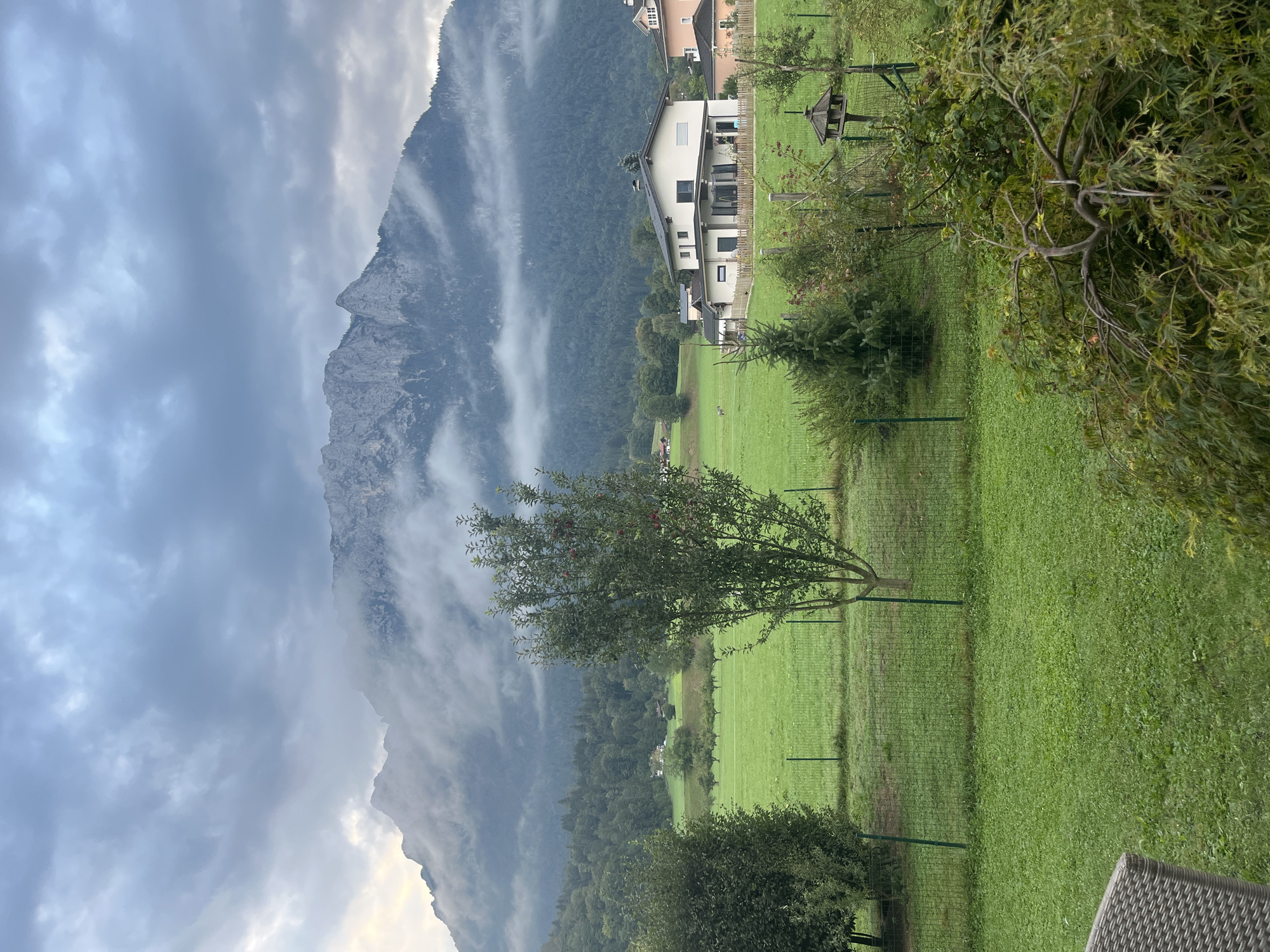 This is a photograph taken in Oberndorf, Austria, on a cloudy day. The scene depicts a rural landscape with a focus on a mountain range in the background, partially obscured by mist and clouds. In the foreground, there is a well-maintained green lawn, bordered by a fence. Several trees are visible, including a mature apple tree with red fruit. Beyond the lawn, there are houses, a field, and a forested area leading up to the mountains. A modern-looking house with a white facade and dark window frames is prominently featured in the middle ground. The sky is overcast with various shades of gray and white clouds, with a hint of pale light suggesting it might be early morning or late afternoon. The overall atmosphere is calm and serene.