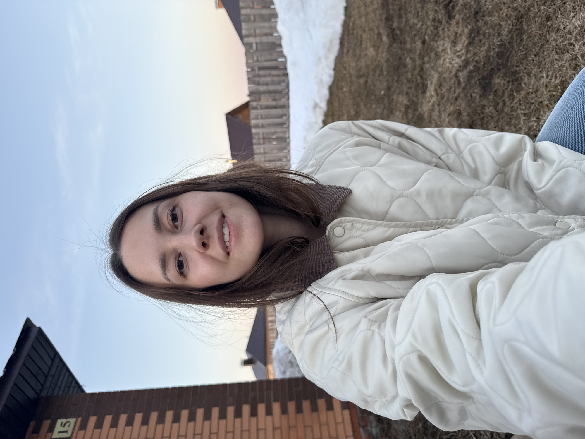 A young woman smiles at the camera while wearing a white quilted jacket. The background shows a brick building with the number "15" visible. Beyond the building, there's a wooden fence and a patch of snow-covered ground. The sky is light blue with hints of dusk or dawn. The location appears to be in a residential area, possibly Chaikovsky, Russia, based on the query.
