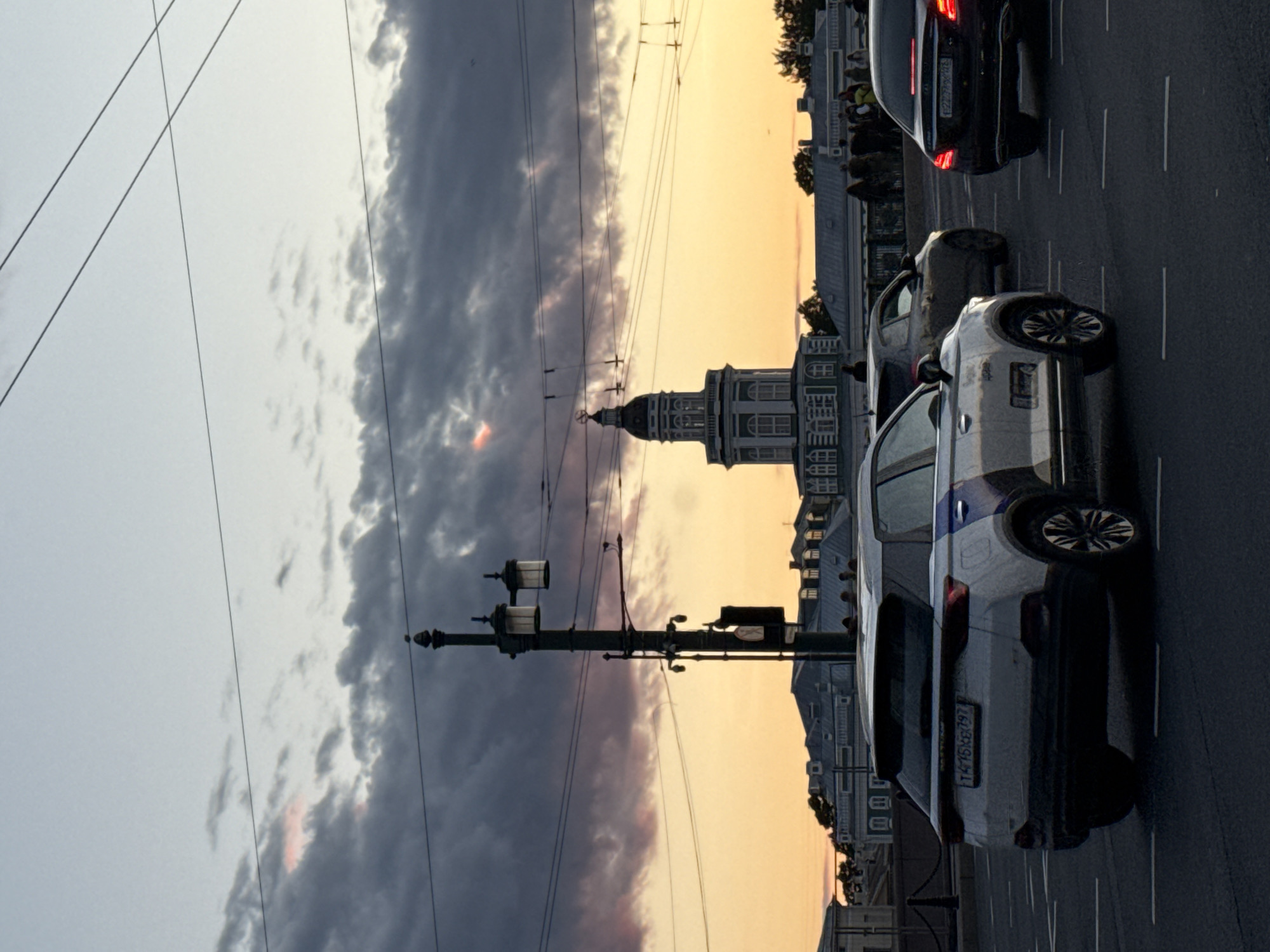 The image captures a street scene in Saint Petersburg, Russia, during the golden hour. In the foreground, several cars are driving on a road. The closest vehicle is a white SUV with blue and black accents, displaying a license plate that reads "T475KX 797". To its right, a black sedan is visible with its brake lights illuminated, bearing a license plate starting with "E223". Further behind the white SUV, a dark gray hatchback is also present.
The middle ground features a street lamp with two lanterns. Beyond that, a distinctive building with a green tower and white architectural details is prominent against the sky. This structure is likely the Kunstkamera Museum, a landmark located on the Spit of Vasilyevsky Island, which aligns with the perspective and architectural style. The background consists of a line of buildings with mansard roofs, indicative of St. Petersburg's architectural heritage, and a distant view of trees and more buildings.
The sky is a mix of dramatic, dark clouds tinged with pink and orange hues from the setting sun, creating a vibrant contrast with the lighter sky above. Several power lines stretch across the frame, common in urban environments. A small group of pedestrians can be seen on a bridge or embankment in the background, suggesting an active public space even as daylight fades. The overall atmosphere is one of urban activity transitioning into evening under a striking twilight sky.