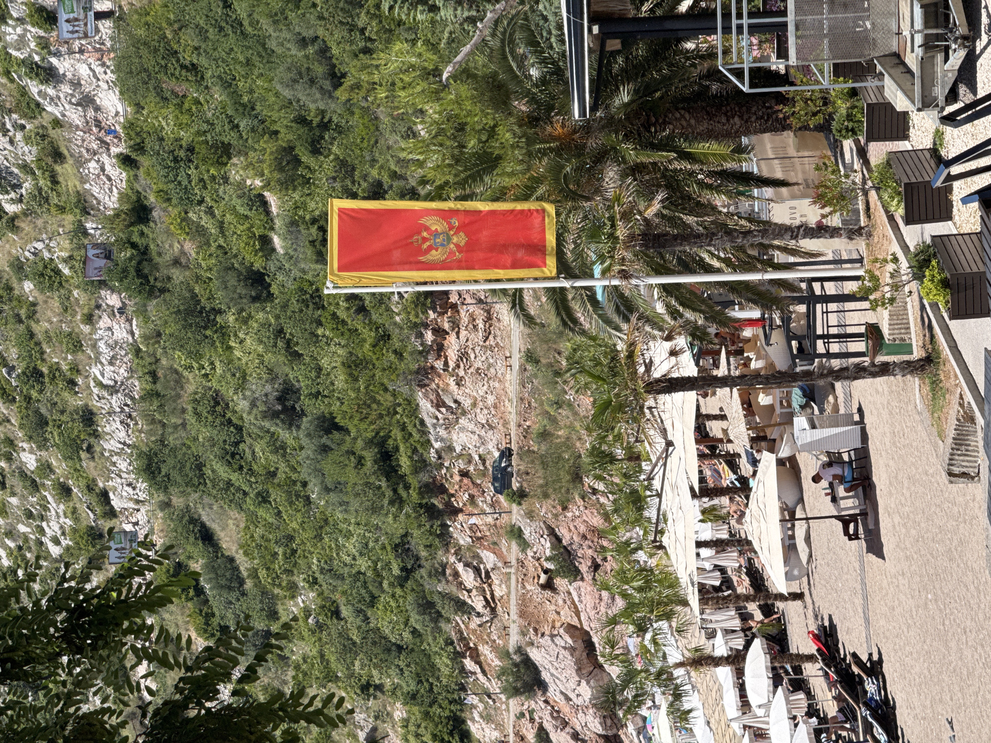 The image shows a view of the beach in Kamenovo, Montenegro. A red and gold Montenegrin flag flies prominently in the foreground. The beach is lined with white umbrellas and lounge chairs, with several people relaxing on the sand. Palm trees are scattered throughout the scene, and a road winds up a rocky, vegetated hillside in the background. A dark-colored car is visible on the road. The overall impression is of a sunny day at a popular tourist destination.