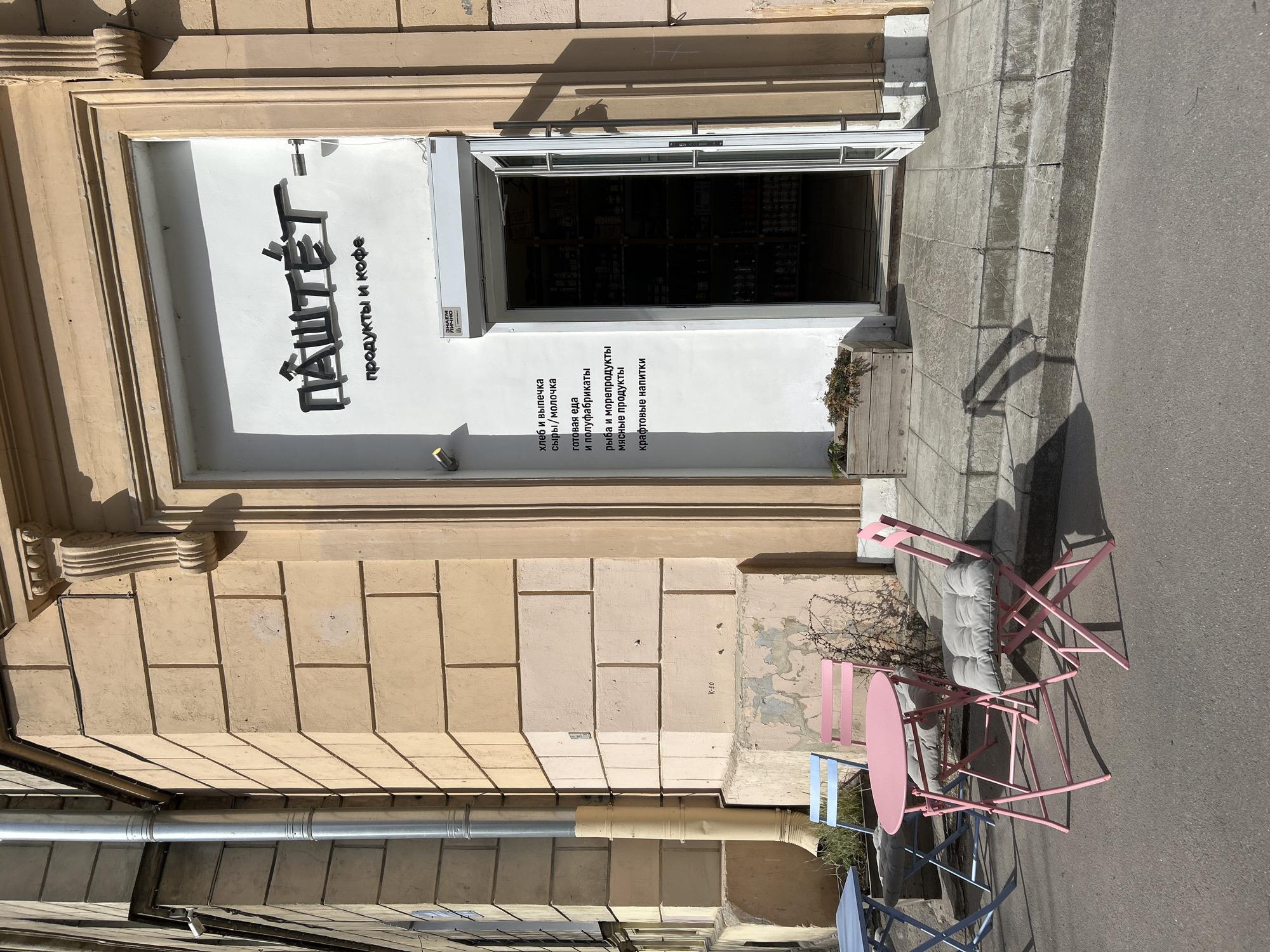 An outdoor, eye-level shot captures the entrance of a grocery and coffee shop named "ПАШТЕТ" (Pâté), located in Saint Petersburg, Russia. The storefront features a white facade with a dark, open doorway, above which an awning or security shutter is visible. The building's exterior is a light, warm tone, possibly stucco or stone blocks, with decorative architectural elements around the doorway.
The shop's name, "ПАШТЕТ-", is prominently displayed in large black block letters above the entrance. To the right, smaller text reads "продукты и кофе" (products and coffee). Below this, a list of offerings is visible: "хлеб и выпечка" (bread and pastries), "сыры/молочка" (cheeses/dairy), "готовая еда и полуфабрикаты" (ready meals and semi-finished products), "рыба и морепродукты" (fish and seafood), "мясные продукты" (meat products), and "крафтовые напитки" (craft drinks). A small label with "ЭНДЫМ ЛИЧНО" is also present near the door. On the building facade, "K-50" is faintly visible.
In front of the shop, on the concrete sidewalk, are two sets of outdoor seating. Closer to the viewer is a pink, round table with two matching pink folding chairs, one with a grey cushion. Further to the left, partially obscured, is another set of blue chairs and a table. A small, dry plant in a wooden planter sits next to the shop's entrance. The scene is brightly lit by the sun, casting strong shadows from the building and furniture, suggesting it is mid-day or afternoon on a clear day. A metal gutter runs down the building's left side. No people are visible.