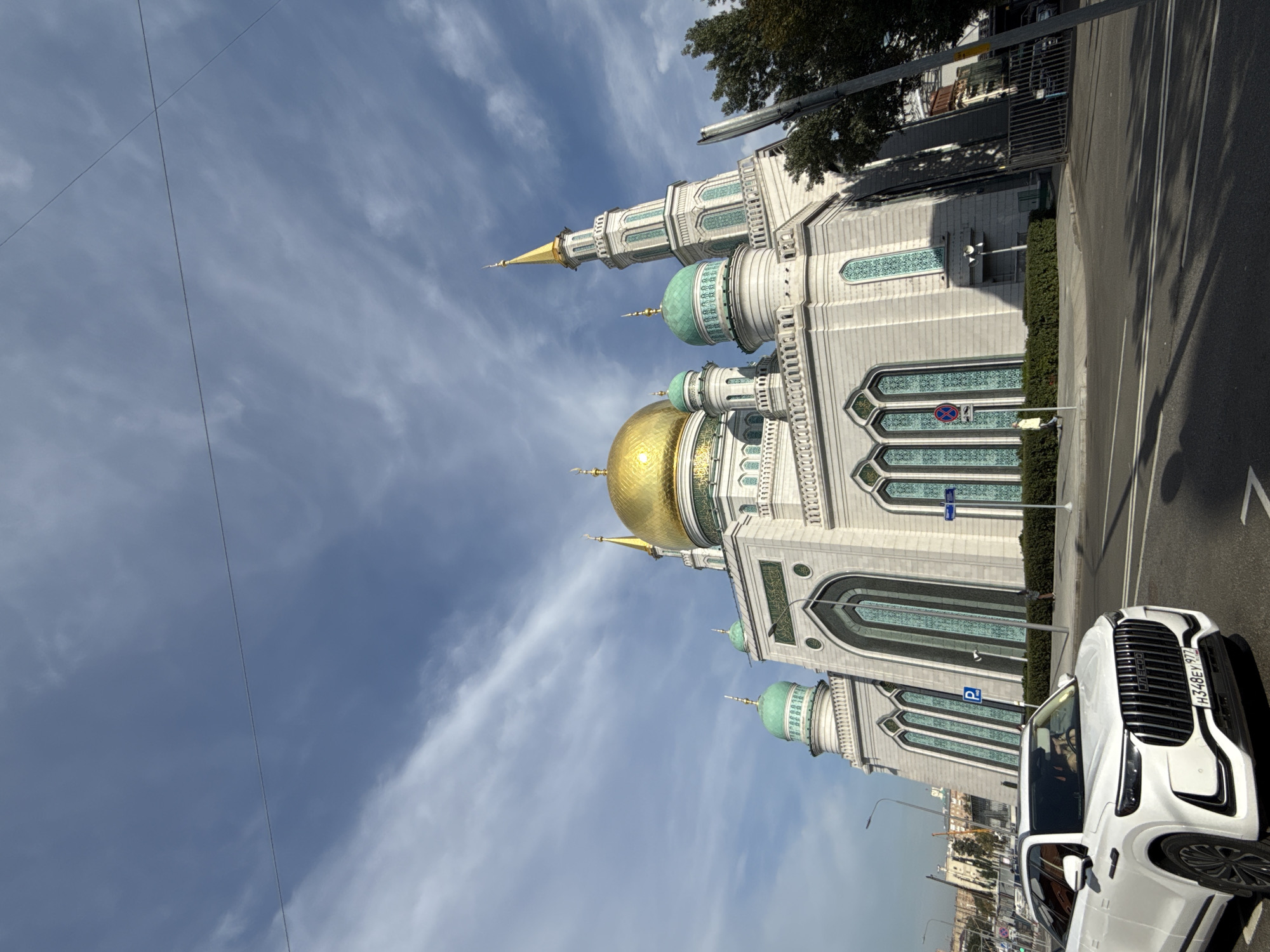 The image shows the Moscow Cathedral Mosque, a large religious building with a golden dome and several turquoise domes, located in Moscow, Russia. The mosque features intricate architectural details, including arched windows with turquoise patterns and decorative friezes. The building is constructed from light-colored stone. In the foreground, a white SUV is parked on the right side of a street. The sky above is blue with scattered clouds, and trees are visible on the right side of the image. The scene appears to be set during the daytime.
