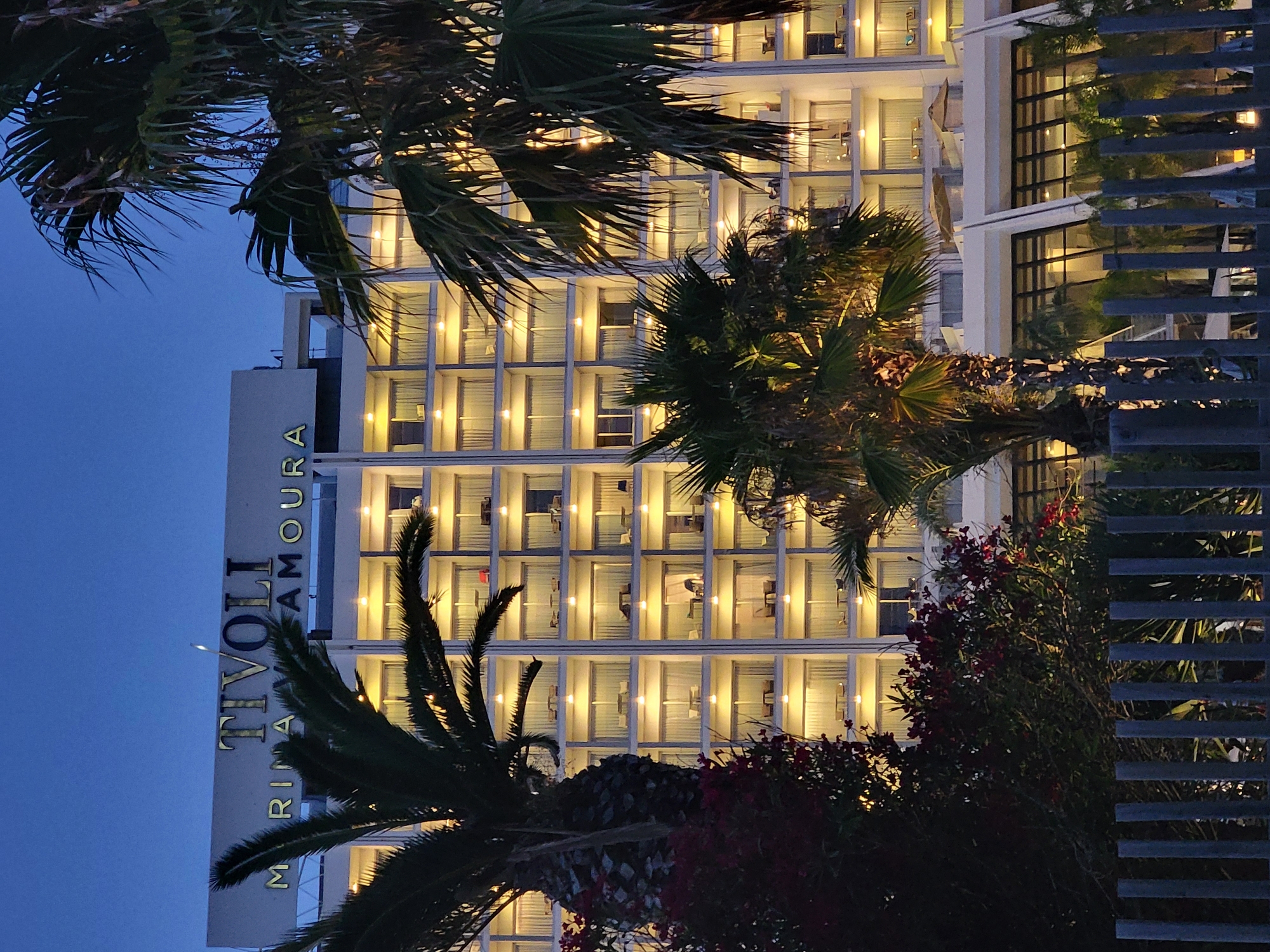 An outdoor, eye-level shot captures the Tivoli Marina Vilamoura hotel at dusk in Quarteira, Portugal. The hotel, a multi-story white building with many illuminated balconies, dominates the frame. Palm trees and dark red flowering bushes are in the foreground, partially obscuring the lower floors of the hotel. A metal fence runs along the bottom of the image. The sky is a deep blue, indicating twilight.