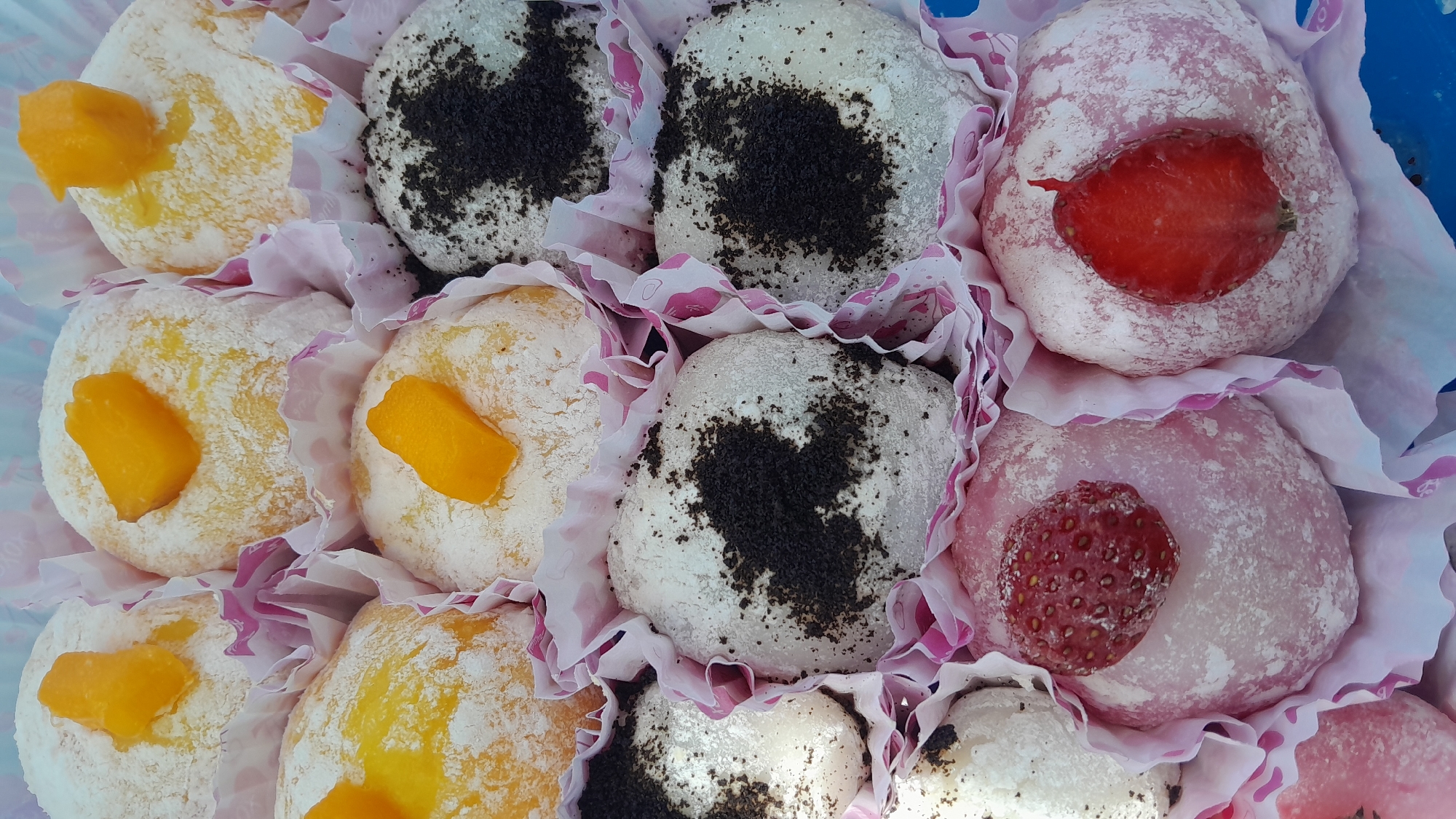 This is a close-up, top-down shot of a box of assorted mochi desserts. The mochi are individually placed in pink and white fluted paper liners. There are at least three varieties visible: some topped with pieces of orange fruit, some covered in dark, crumbly topping that resembles crushed cookies, and some pink mochi topped with fresh strawberries, either whole or halved. The mochi themselves appear to be dusted with white powder, likely confectioners' sugar or rice flour. The lighting is bright and even, suggesting an indoor setting, possibly a food stall or a bakery. Based on the visual cues, this scene could be located in a market or a dessert shop in Bandung City, Indonesia.