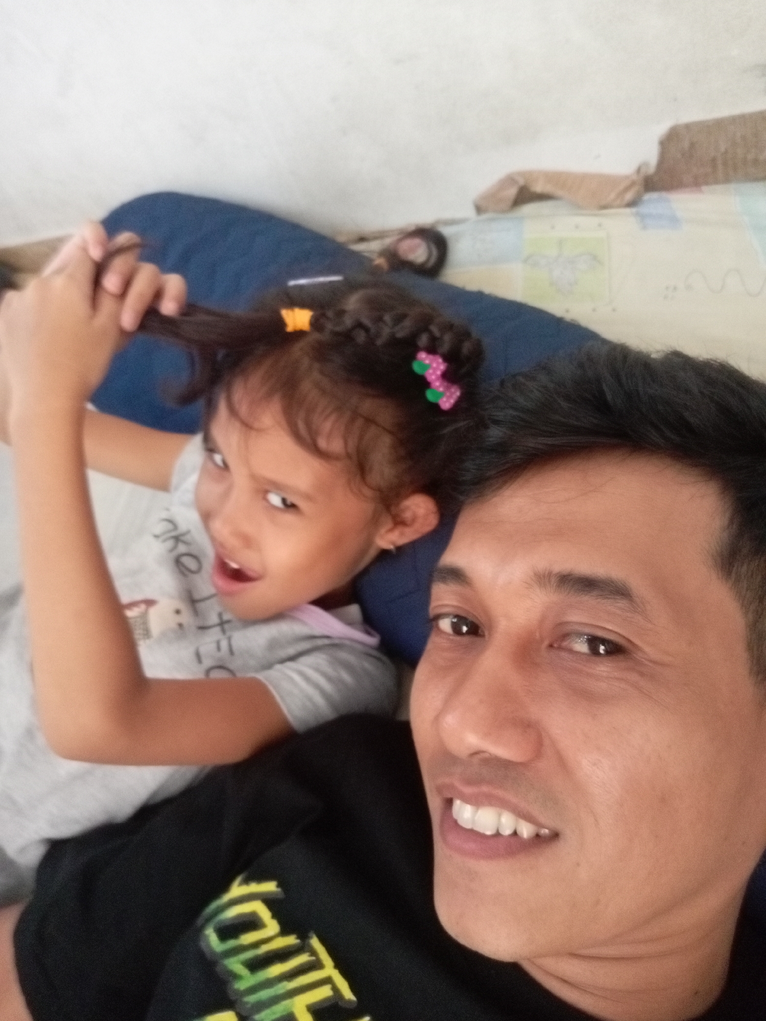 This media file is a selfie taken by a man and a young girl, likely father and daughter, lying down together. The man is in the foreground, smiling and looking at the camera. The girl is behind him, with her arm raised, playfully pulling on her braided hair, which is decorated with colorful barrettes. She is looking off to the side with an open mouth, perhaps in mid-sentence or mid-laugh. Both are wearing casual clothing. The background appears to be an indoor setting, possibly a bedroom or living area, with a blue cushion and patterned bedding visible. The overall mood is affectionate and lighthearted. There is no explicit indication of the time of day or weather conditions. A partial inscription is visible on the girl's grey t-shirt, reading "nke". There is also visible text on the man's black shirt. The scene suggests a relaxed moment shared between the two individuals.