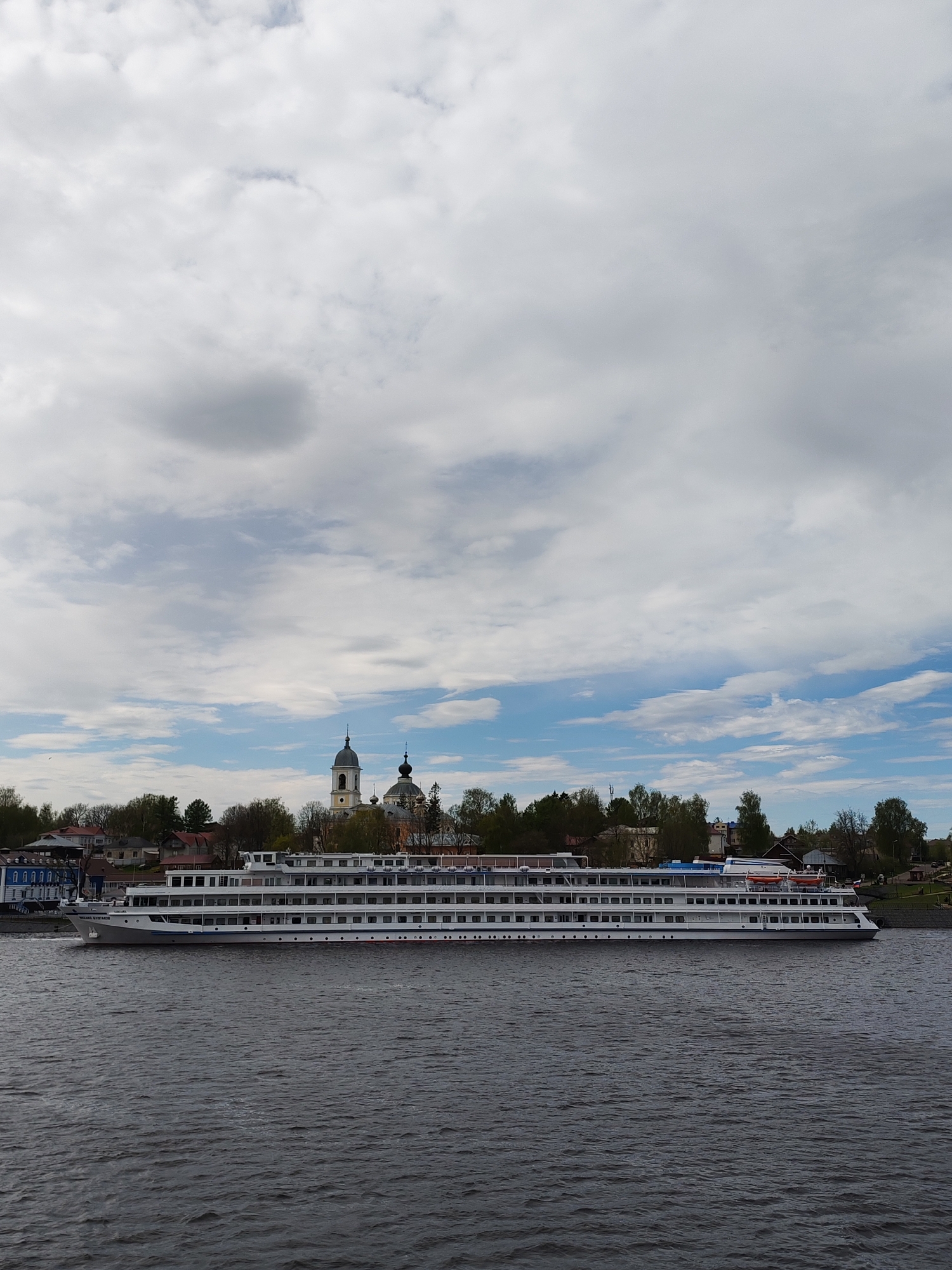 A long, white passenger cruise ship is on the water in front of Myshkin, Russia. The ship has multiple decks and appears to be docked or moving very slowly along the river. The skyline of Myshkin is visible in the background, with a church featuring a tall, light-colored bell tower and dark-colored domes standing out amidst the trees and other buildings. The sky is filled with a mix of white and grey clouds. The water is dark, with slight ripples on the surface.