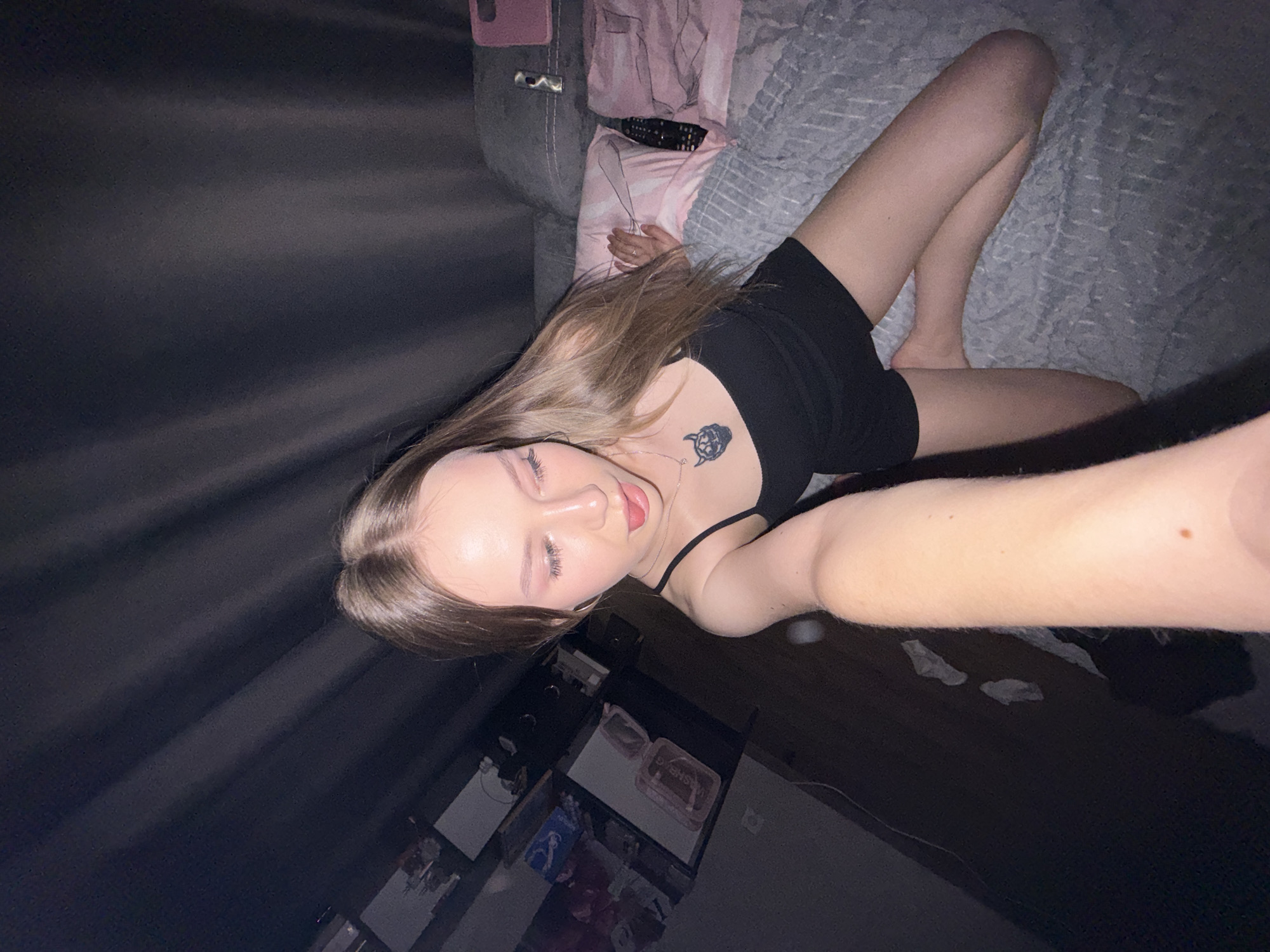A young woman lies on a bed in a dimly lit room. She is wearing a black spaghetti-strap dress and has a tattoo on her upper chest. The bed is covered with a gray blanket. A gray sofa is visible next to the bed, with a pink blanket and a remote control on it. In the lower portion of the image, there is a cabinet or dresser with several items on it. It is not possible to verify the location is Chaikovsky, Russia.