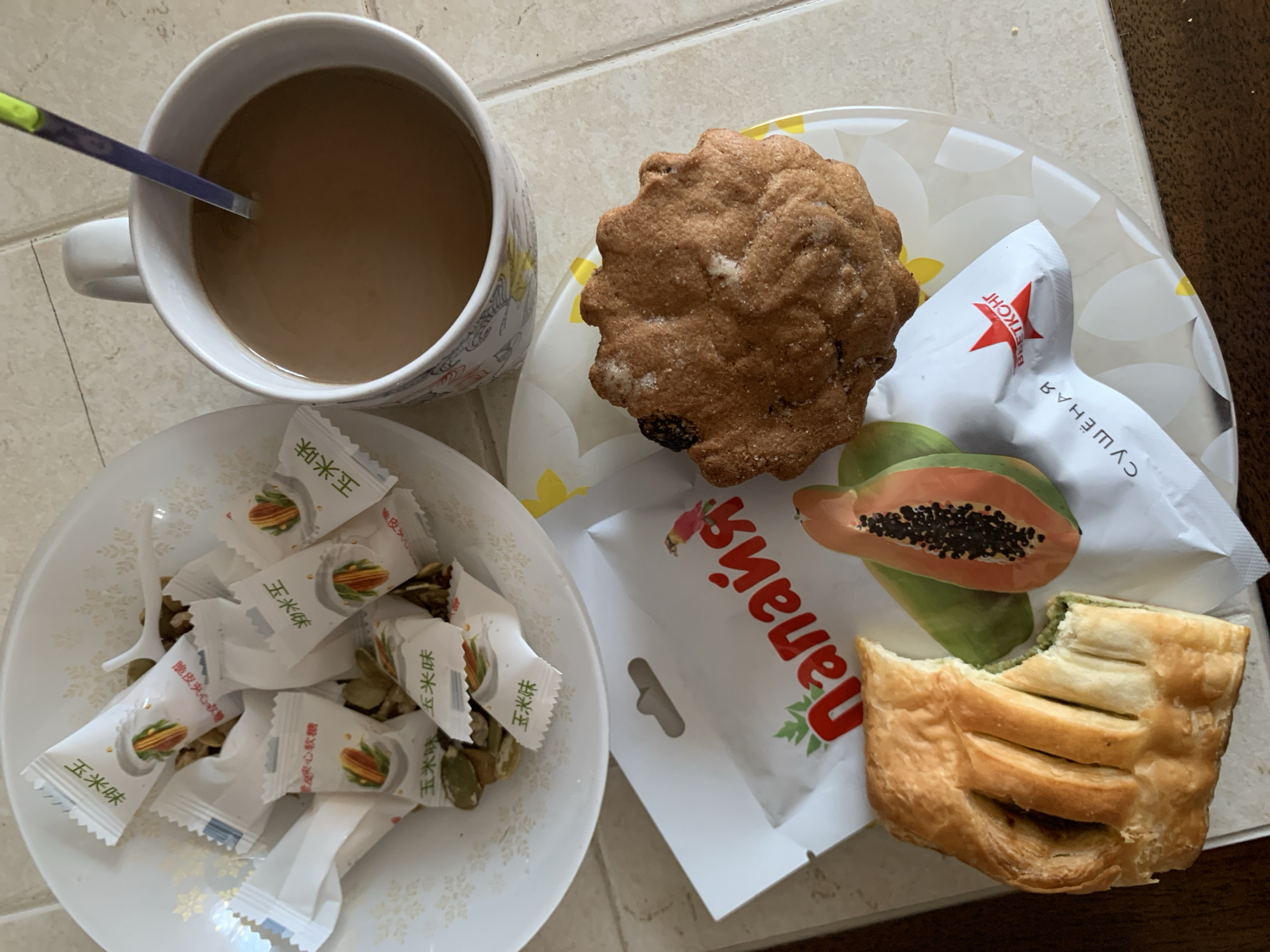 A high-angle, slightly tilted full shot shows a breakfast spread on a tiled surface. In the top left, a white bowl contains numerous individually wrapped candies, likely with a corn flavor based on the packaging. Scattered among the candies are pumpkin seeds. To the right, a white ceramic mug with a decorative rim holds a brown beverage, presumably coffee or tea, stirred by a blue and yellow utensil. In the foreground, a white plate holds a muffin and a pastry. The muffin is golden brown with a textured surface, sprinkled with sugar. The pastry, partially eaten, is a flaky, golden-brown dough with visible green filling. Next to these items is a slice of papaya, showing its vibrant orange flesh and black seeds. A white paper bag with red lettering and a bird illustration is also present, along with another white packet featuring a red star logo and text. The overall scene depicts a simple breakfast, likely at home. The specific location of Kapyĺ, Belarus is not discernible from the visual information in the image.
