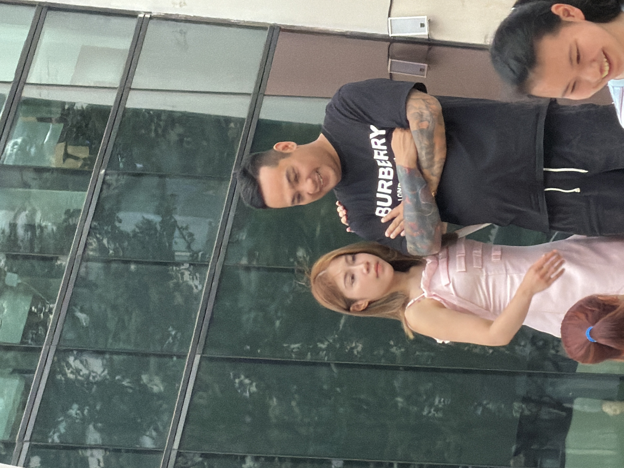 An Asian man and an Asian woman are standing in front of a building with a glass facade in Yuzhong District, China. The man is wearing a black t-shirt with "BURBERRY LONDON" printed on it, and he has tattoos on his arms. He is smiling and has his arms crossed. The woman is wearing a pink satin dress with bows. She is looking to the side. In the foreground, the back of a young Asian girl's head is visible. She has dark hair tied in a ponytail. The building's facade is made of green tinted glass panels framed by metal. The overall scene appears to be outdoors during the daytime.
