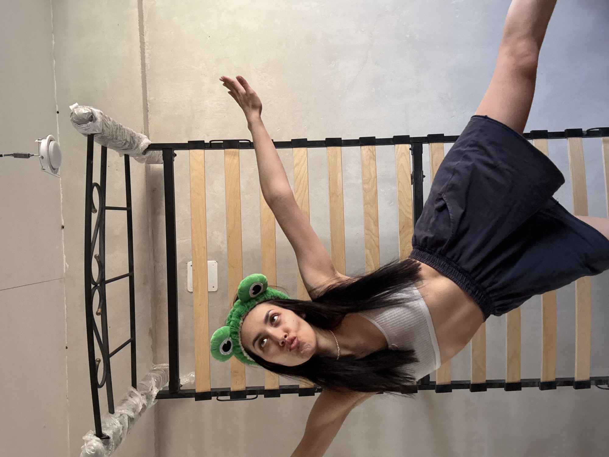 A young woman wearing a white ribbed tank top, dark shorts, and a green frog-themed hat is oriented diagonally in the frame. She has long dark hair and is making a kissing face towards the camera. She has one arm raised and one arm extended diagonally downward.
She is positioned in front of an unassembled bed frame. The headboard of the bed is wrapped in clear plastic. The environment appears to be an unfinished room with bare walls and a simple light fixture. The location is not discernable as Votkinsk, Russia.