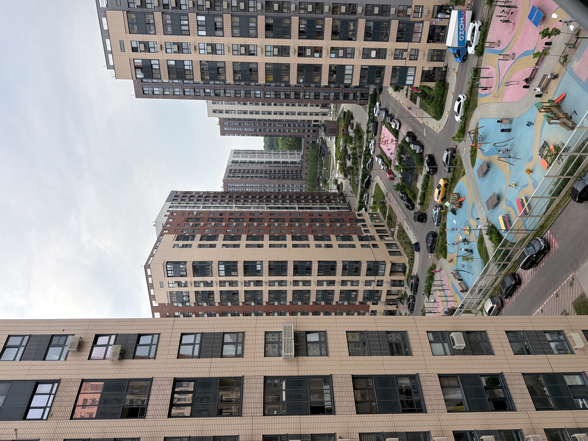 This is an aerial view of a modern residential complex in Moscow, Russia, likely in a newly developed area given the contemporary architecture and the presence of a children's playground. The scene is dominated by several tall, multi-story apartment buildings constructed with a combination of light-colored brick, darker paneling, and numerous windows. In the foreground, the side of a light tan brick building is visible, featuring rows of dark-framed windows, some with air conditioning units mounted externally.
In the middle ground, a vibrant children's playground with colorful rubberized surfacing in shades of blue and pink is a central feature. It includes various play structures such as slides, swings, climbing frames, and benches. Several adults and children are present, engaged in recreational activities. A road network surrounds the playground, with numerous cars parked and driving. A white delivery truck with "OZON" branding is visible.
In the background, more apartment buildings of similar modern design are scattered across the landscape, interspersed with green spaces and trees, suggesting a well-planned urban environment. The sky is overcast with a pale, diffused light. The overall impression is one of a densely populated but functional urban residential area.