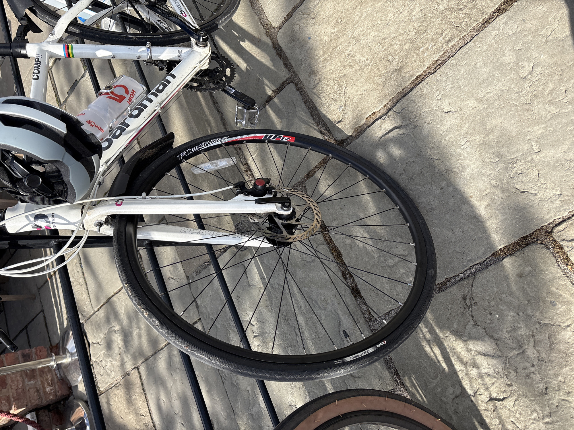 The image depicts a close-up view of a white road bike, with a white helmet leaning against the frame. The bike is equipped with disc brakes and black tires. A water bottle is attached to the frame. The bike is resting on a metal rack or stand. The background consists of light gray paving stones with visible cracks and variations in texture. Another bike is visible in parts in the frame with tan-colored tires, seemingly stacked behind the main bike. The location is most likely outdoors on a paved surface. Trafford, United Kingdom is not discernible based on visual evidence within the image.