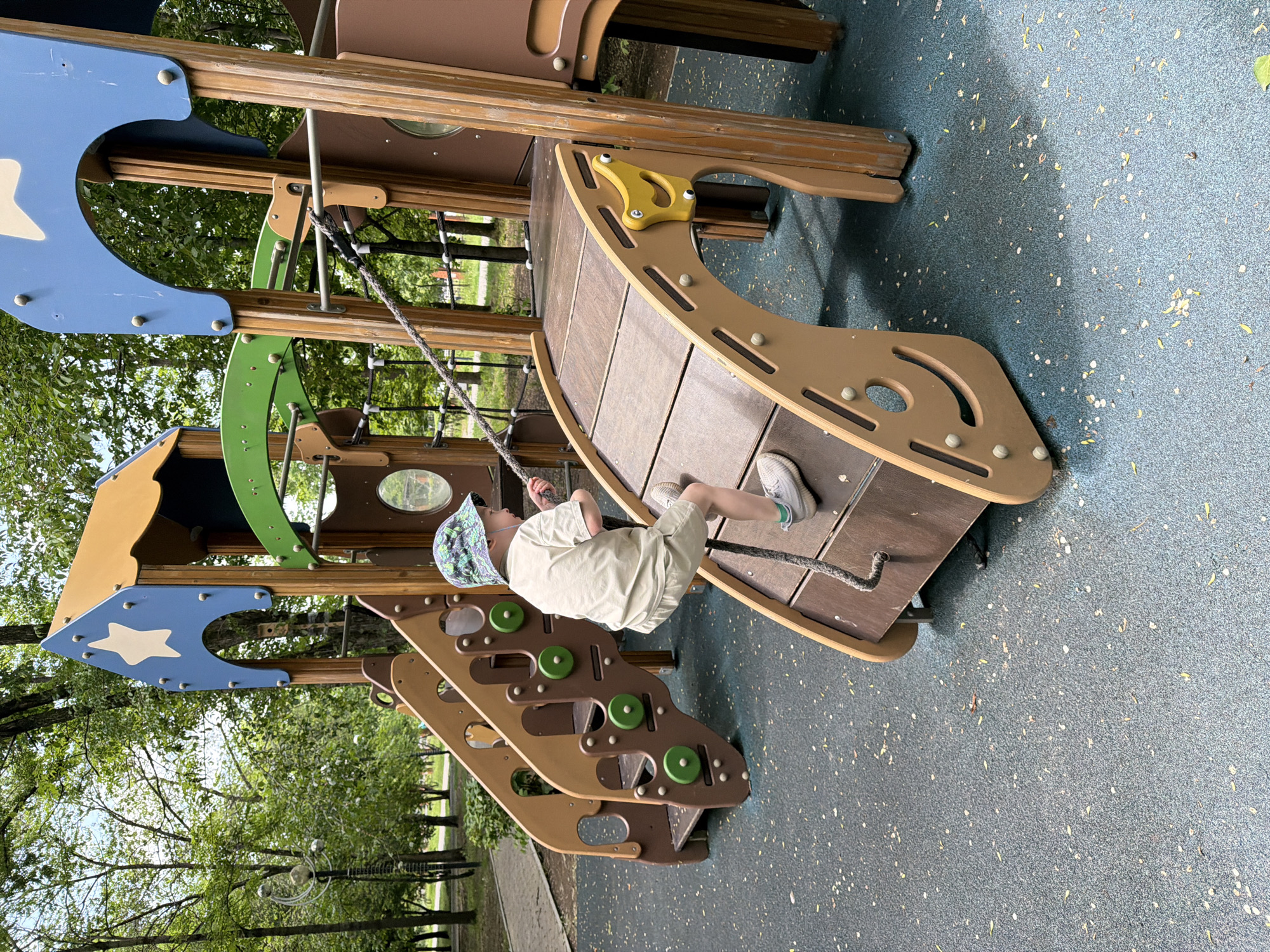 A young boy wearing a light-colored t-shirt, light-colored shorts, and a patterned bucket hat is climbing a rope on a wooden and plastic playground structure. The playground has slides and climbing elements. The boy is on the right side of the image, facing away from the camera and to the left. He is positioned on a sloped wooden platform, holding onto a thick rope. The playground surface is a speckled blue rubber mat. In the background, there are trees and foliage, suggesting an outdoor park setting. The overall scene depicts a child engaged in play on a playground.