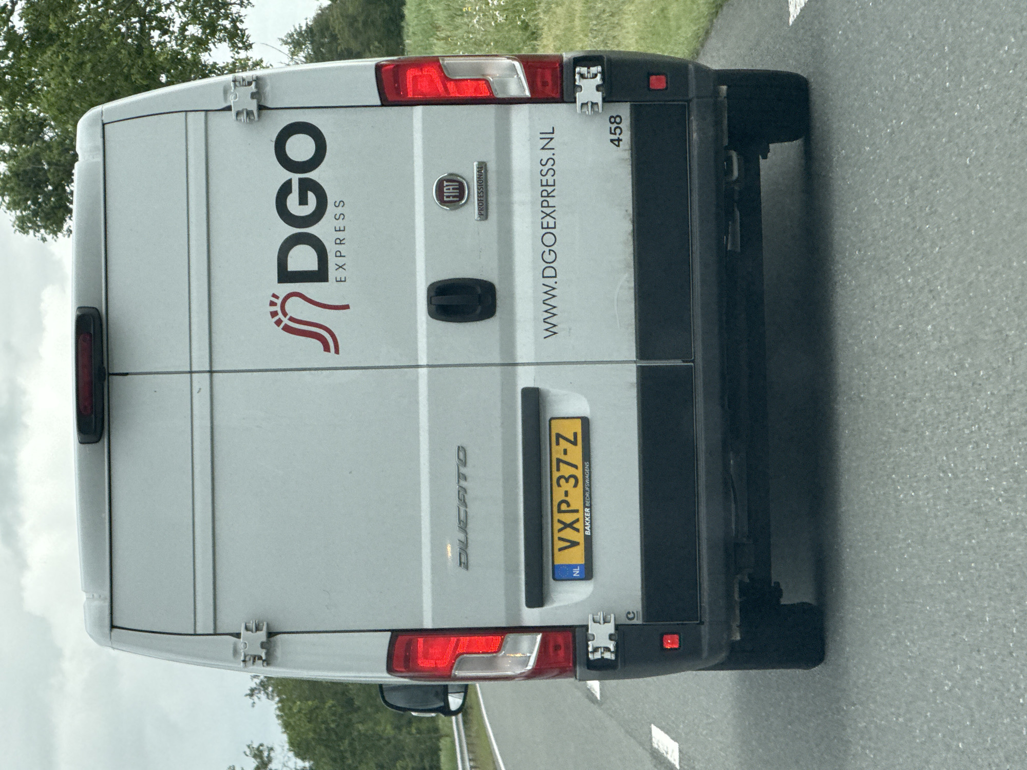 The image shows the rear of a white DGO Express van. The van has a license plate that reads "VXP-37-Z" and has "NL" to the left of it. Below the license plate, it says "BAKKER BEDRIJFSWAGENS". The van also has "DUCATTO" written on the back, as well as the Fiat logo and the word "PROFESSIONAL". On the right side of the van, the website "WWW.DGOEXPRESS.NL" is visible, along with the number "458". The van is on a paved road, possibly a highway, with a grassy shoulder and trees in the background. The sky is cloudy. The vehicle is positioned on the right lane of the road. No people are visible. The scene appears to be a daytime shot. The location of Leeuwarden, Netherlands is not explicitly evident from the image.