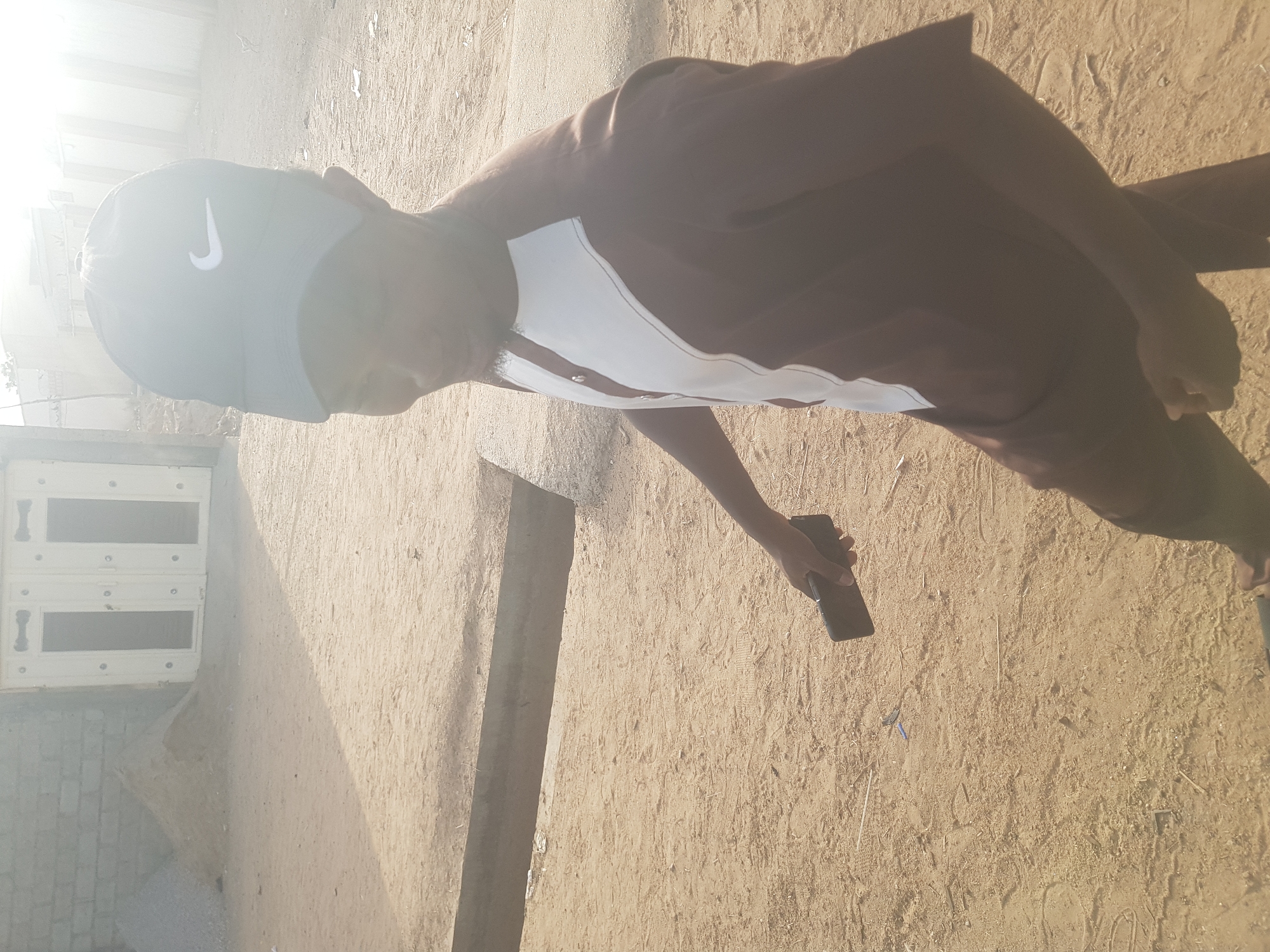A person wearing a Nike baseball cap, a brown and white shirt, and dark pants is standing outdoors in Dabawa, Nigeria. The person is holding a black mobile phone in their left hand. The ground is sandy and dusty. In the background, there is a white building with a door and a partially visible wall. The lighting is bright, and the image appears to have been taken during the day.