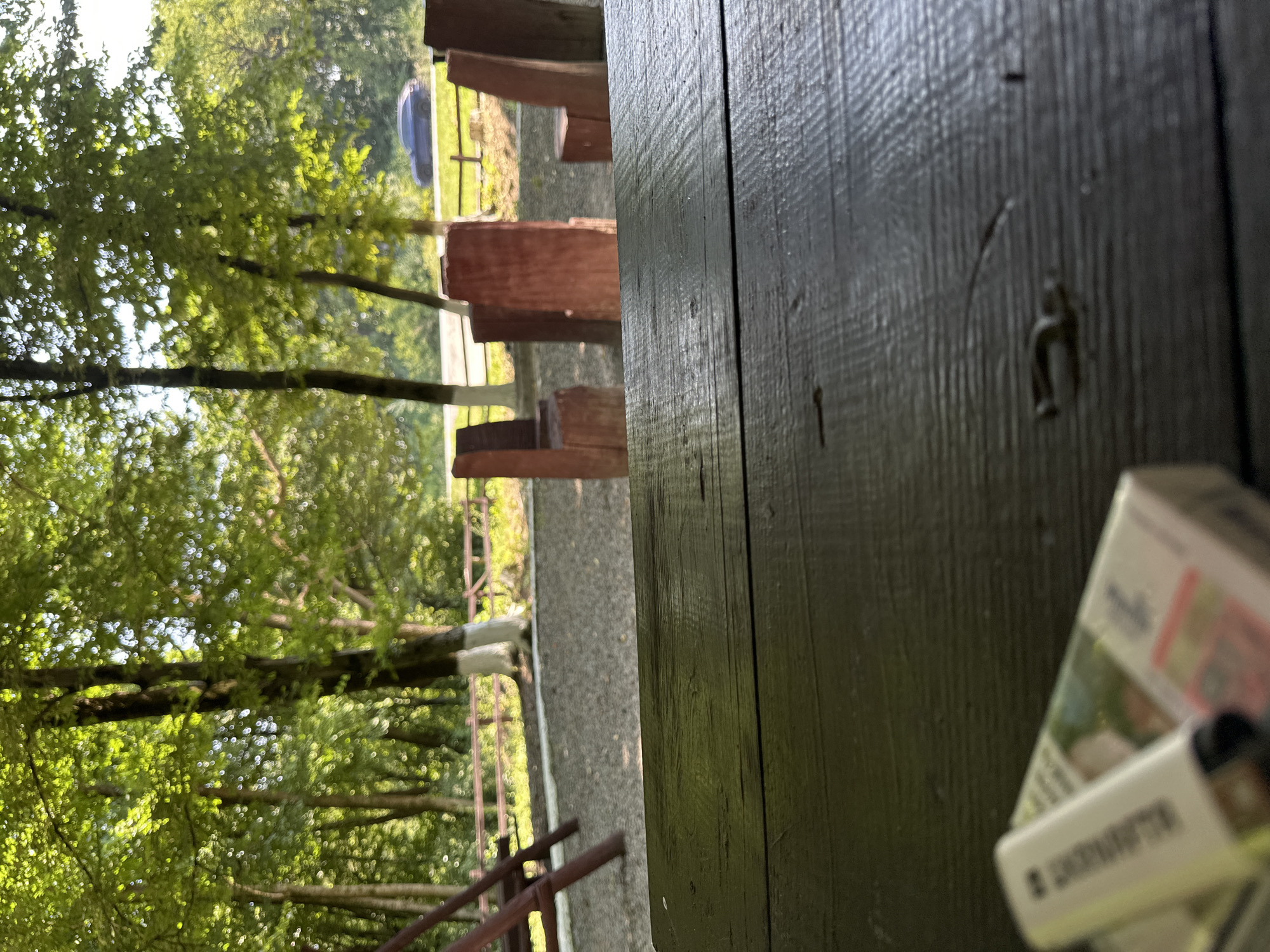 The image depicts a wooden table in the foreground, with a cigarette pack and a lighter on it. In the background, there is a park-like setting with trees, a gravel path, wooden benches, and a road with a car visible. The trees are green and lush, suggesting a warm season. The overall impression is of an outdoor recreational area, possibly a park or picnic spot. There are no people visible in the image. The location is identified as Shatava, Ukraine.