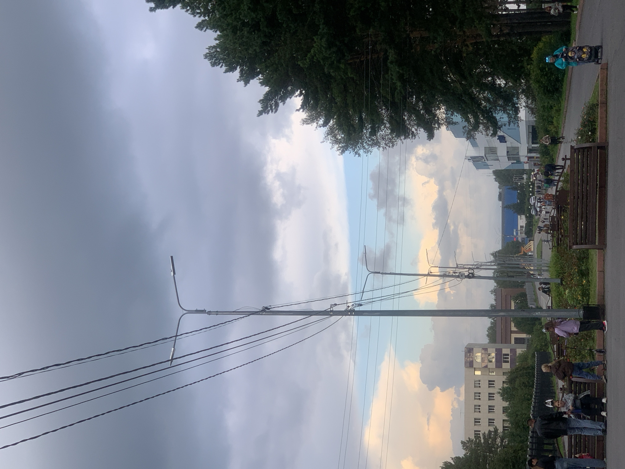 The image depicts a street scene in Prokopyevsk, Russia, likely on a cloudy day with some patches of blue sky visible. In the foreground, several people are standing and sitting on benches. A woman is pushing a stroller with a child in it. Power lines stretch across the sky, supported by utility poles. A street lamp with two lights is prominent on the right side of the image.
In the midground, there is a paved road with more people walking. Buildings line the street, including a light-colored multi-story building on the left and a modern blue and white building on the right. Trees, including tall evergreens, are visible along the roadside. A more distant view shows more buildings and infrastructure, suggesting an urban or suburban environment. The ground in the foreground appears to be a sidewalk or pedestrian area with benches and some greenery.
