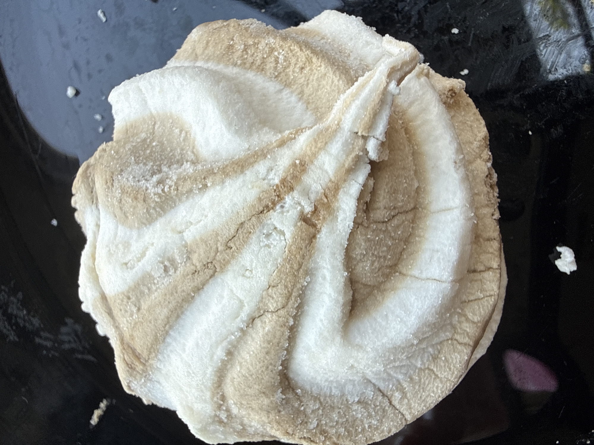 A swirl-patterned meringue sits on a black surface. The meringue is mostly white with streaks of light brown. The surface is dark and appears to be a plate or tray. There are a few white specks scattered on the black surface around the meringue.