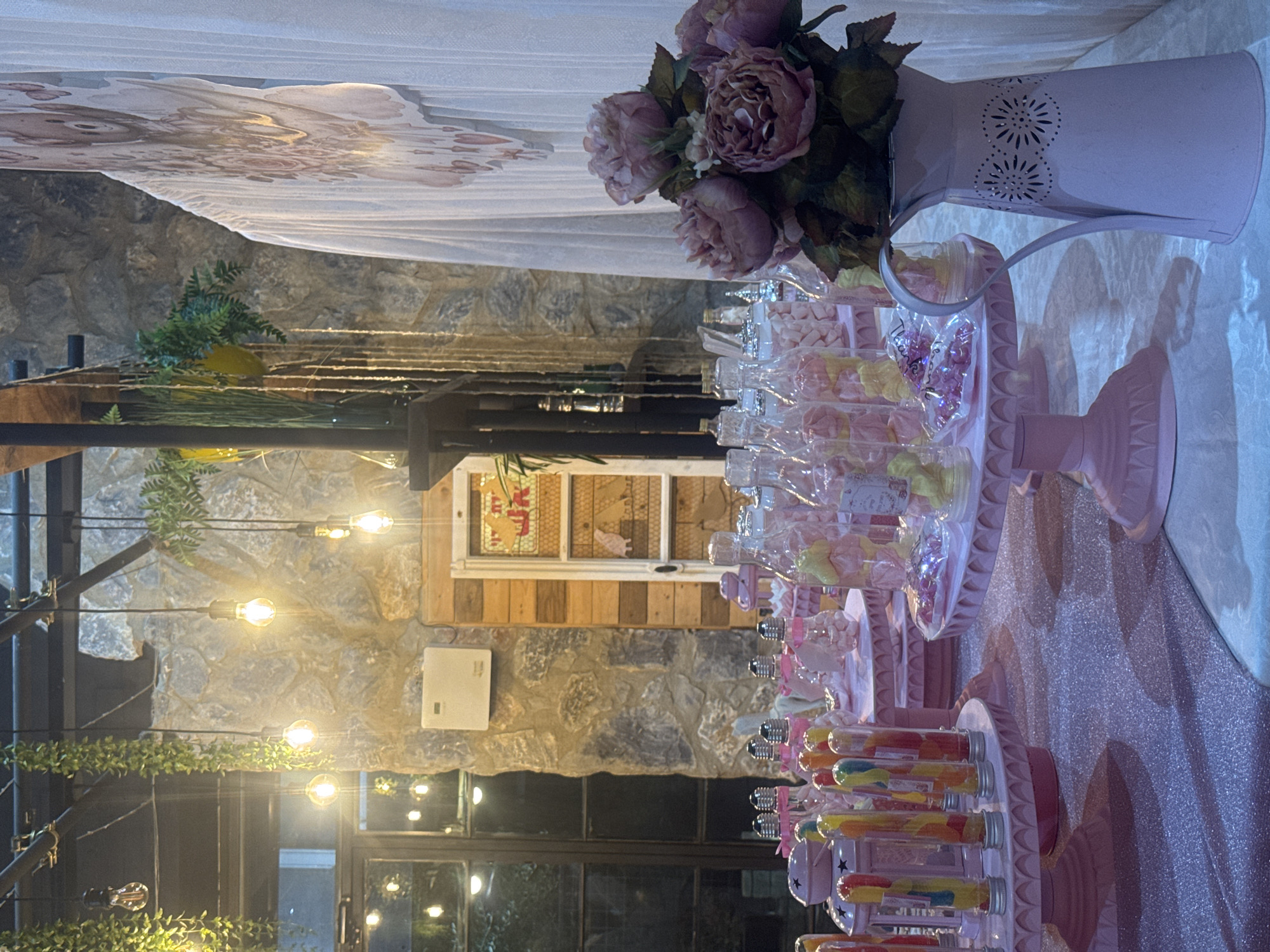 This image depicts a dessert table setup, likely for a party or celebration, in what appears to be an outdoor or semi-outdoor venue in Migdal HaEmek, Israel. The setting is characterized by a stone wall and wooden elements, possibly part of a building's facade or a decorative structure. Several exposed filament light bulbs hang from above, creating a warm, ambient glow, further enhanced by decorative hanging greenery.

The table itself is covered in a sparkly pink tablecloth and is laden with various sweets. These include candy-filled tubes displayed on pink cake stands, jars filled with colorful candies and marshmallows, and other decorative confectionery items. In the foreground, a light pink metal pitcher holds a bouquet of artificial pink roses and greenery. A white sheer curtain with a patterned overlay, possibly a backdrop, is visible on the right side of the image. The overall aesthetic is whimsical and festive, with a predominant pink color scheme. No people are visible in the image.
