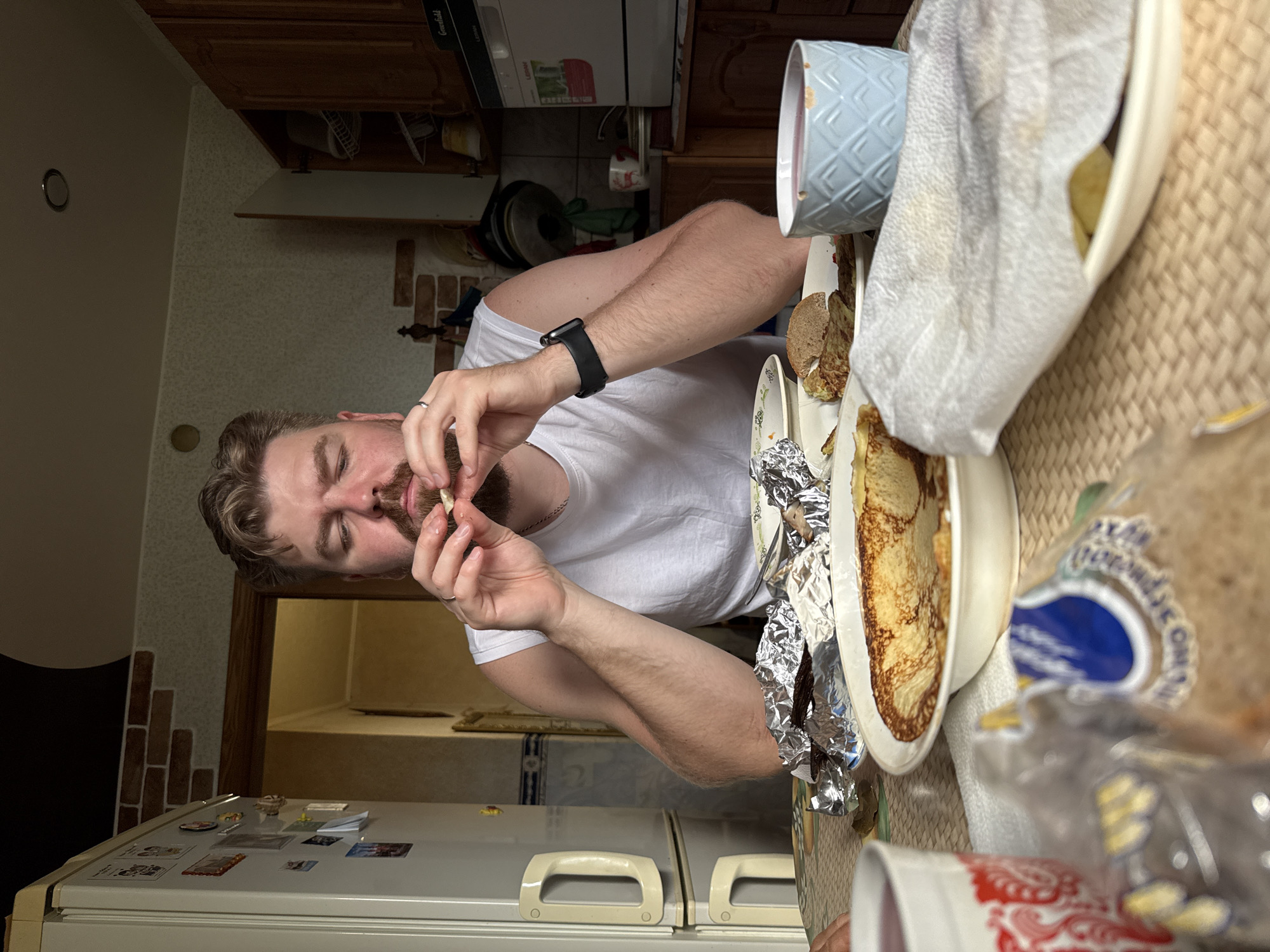 A man with light brown hair and a beard, wearing a white tank top, sits at a table in what appears to be a kitchen. He is holding a piece of food to his face, with his eyes closed. On the table in front of him are several plates with food, including pancakes and bread. There is also a blue cup filled with a pink liquid, and a red and white cup. In the background, there is a refrigerator, kitchen cabinets, and a doorway leading to another room. The overall setting suggests a casual meal.