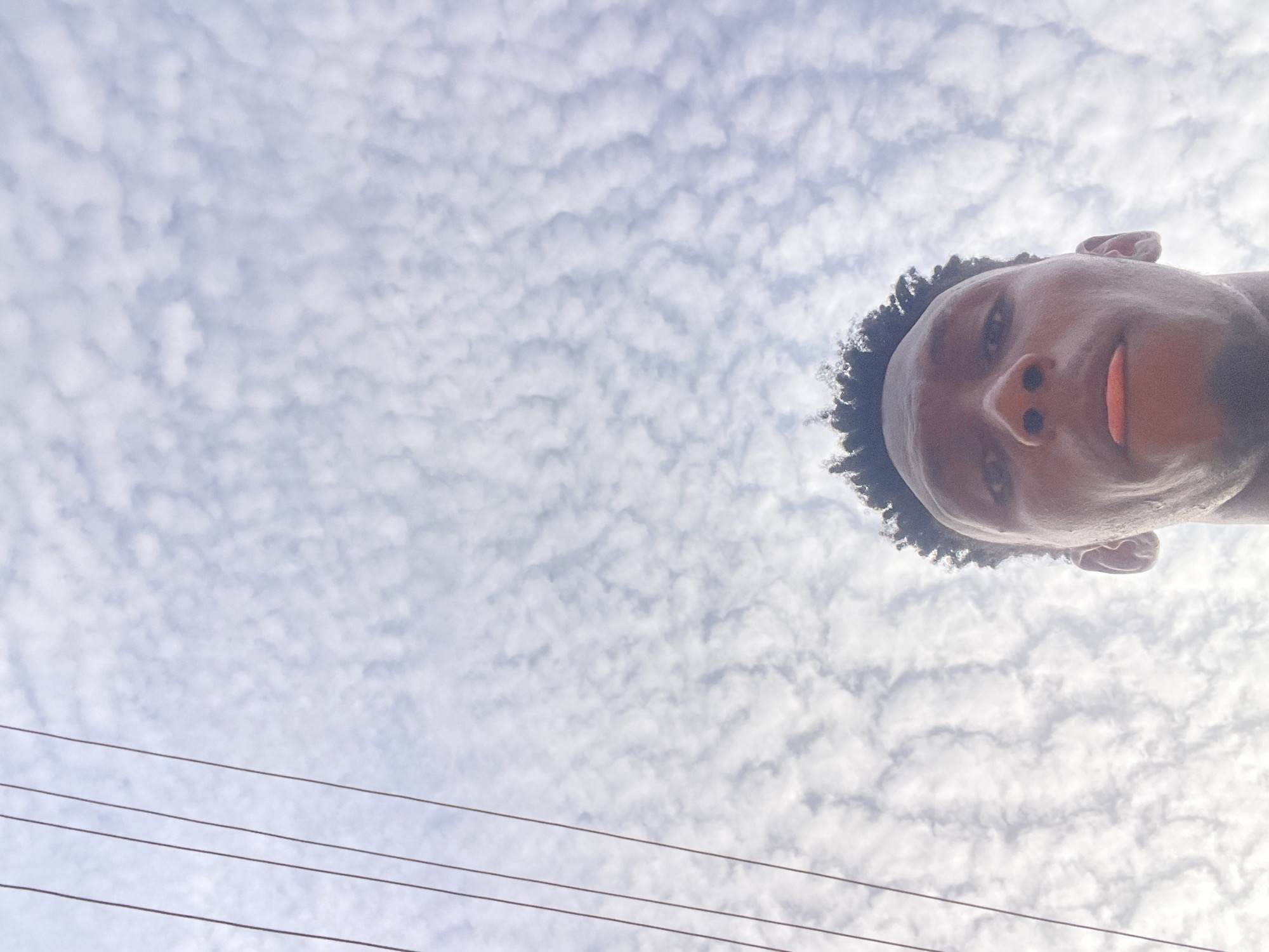The image presents a low-angle view looking up towards the sky, dominated by a cloud-filled expanse. The head of a person, likely a young male with dark skin and short, curly hair, is visible in the upper right portion of the frame. He looks directly at the camera with a neutral expression, and the tip of his tongue is slightly visible.
The sky is largely covered by a dense pattern of small, white, ripple-like clouds, characteristic of altocumulus clouds or a "mackerel sky," suggesting a bright daytime setting. Three dark, parallel utility wires or power lines stretch across the lower left quadrant of the image, contrasting against the bright sky. The composition emphasizes the expansive sky and clouds, with the person and power lines serving as foreground elements from a ground-level perspective. No text is visible. The scene depicts an outdoor environment, with the presence of utility infrastructure. The location context provided is Lokoja, Nigeria.