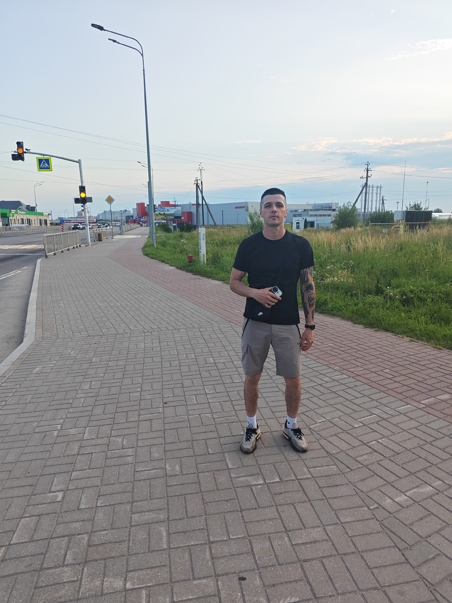A man stands on a paved sidewalk with a brick pattern. He is wearing a black t-shirt, grey cargo shorts, white socks with black stripes, and athletic shoes. He has a black fanny pack around his waist and is holding a small electronic device in his right hand. He has tattoos on his left arm. The background shows a road with traffic, a pedestrian crossing, traffic lights, and streetlights. Beyond that, there are buildings, industrial structures, and a field of tall grass. The sky is light blue with some clouds, suggesting it is daytime, possibly late afternoon or early evening. The scene appears to be in an urban or semi-urban environment. There are some signs visible, but the text is not clearly legible. The man has a neutral expression.