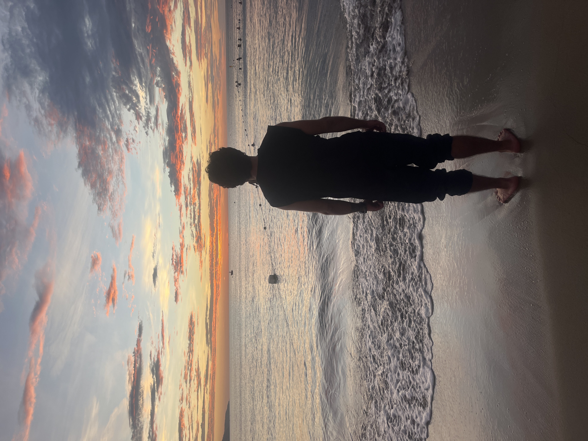 The image captures a tranquil sunset scene on a beach in Đà Nẵng, Vietnam. A person, viewed from behind, stands at the water's edge, facing out towards the ocean and the setting sun. The individual appears to be male, with dark, curly hair, wearing a black t-shirt and dark pants. Their bare feet are partially submerged in the shallow water or wet sand where small waves are breaking.
The sky is a spectacle of vibrant colors, characteristic of a sunset. Rich oranges, yellows, and reds illuminate scattered clouds, contrasting with softer blue and gray tones. These warm hues are reflected on the calm surface of the ocean, stretching towards the horizon. Gentle, foamy waves lap at the sandy shore, creating a soft white line across the wet sand. In the distance, several dark, indistinct objects, possibly buoys or distant boats, are visible on the water, silhouetted against the bright horizon.
The overall atmosphere is one of peaceful observation and appreciation of nature's beauty, as the person stands still, gazing at the expansive view. The time of day is clearly sunset, highlighted by the dramatic sky. There is no visible text in the image.