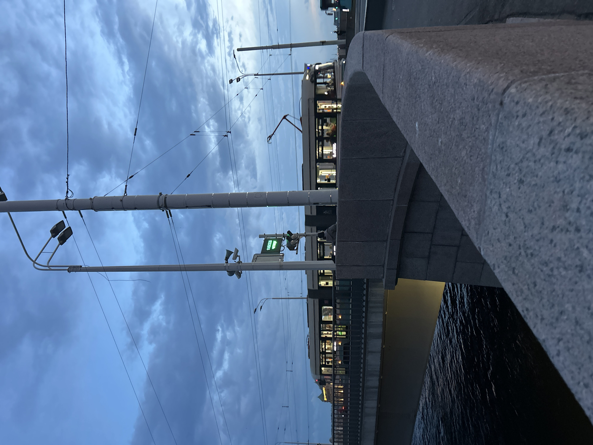 The image depicts a tram moving along a bridge over a dark body of water. The sky is overcast with a cloudy, twilight atmosphere. Streetlights and overhead power lines are visible, along with the illuminated side of the tram showing passengers inside. A stone railing lines the bridge, and in the foreground, there is a stone structure. The scene appears to be an urban environment in the evening.