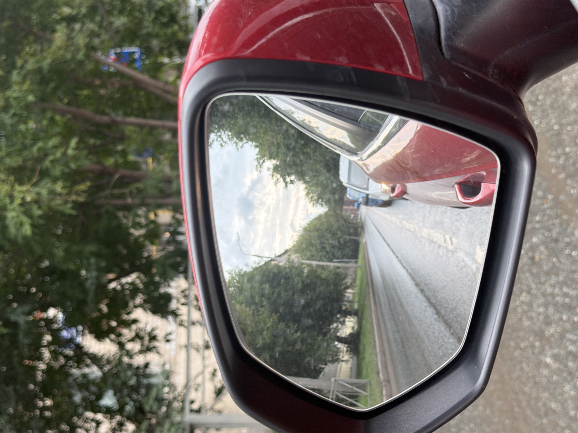 The image captures a reflection in the side mirror of a red car, likely in Kazan, Russia, given the stylistic details and general urban environment. The mirror shows a wet road with visible tire tracks, suggesting recent rain. A line of cars is stopped or moving slowly on the road, with the most prominent car in the foreground of the reflection being a red vehicle, likely the same one the mirror is attached to. Further down the road, several other cars are visible, including a white van and a blue car. Trees line the side of the road, and some buildings are partially visible through the foliage. The sky is cloudy. The focus is on the reflection, with the exterior of the red car partially visible in the foreground. There are no people clearly visible in the reflection or the foreground. The overall impression is a typical urban scene during or immediately after rainfall.