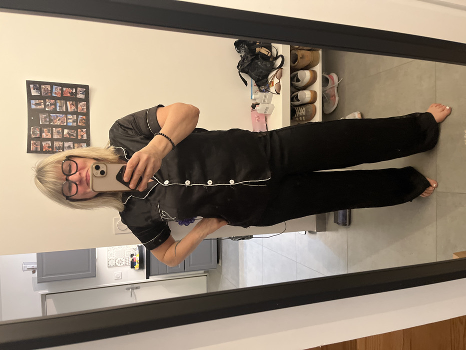 A woman with short blonde hair and glasses poses in front of a mirror, holding a smartphone. She wears black pajamas with white piping. The mirror reflects part of a kitchen with grey cabinets and a refrigerator. A shelf filled with shoes, sunglasses, and other items is visible. A framed photo collage hangs on the wall. The floor is tiled in grey.