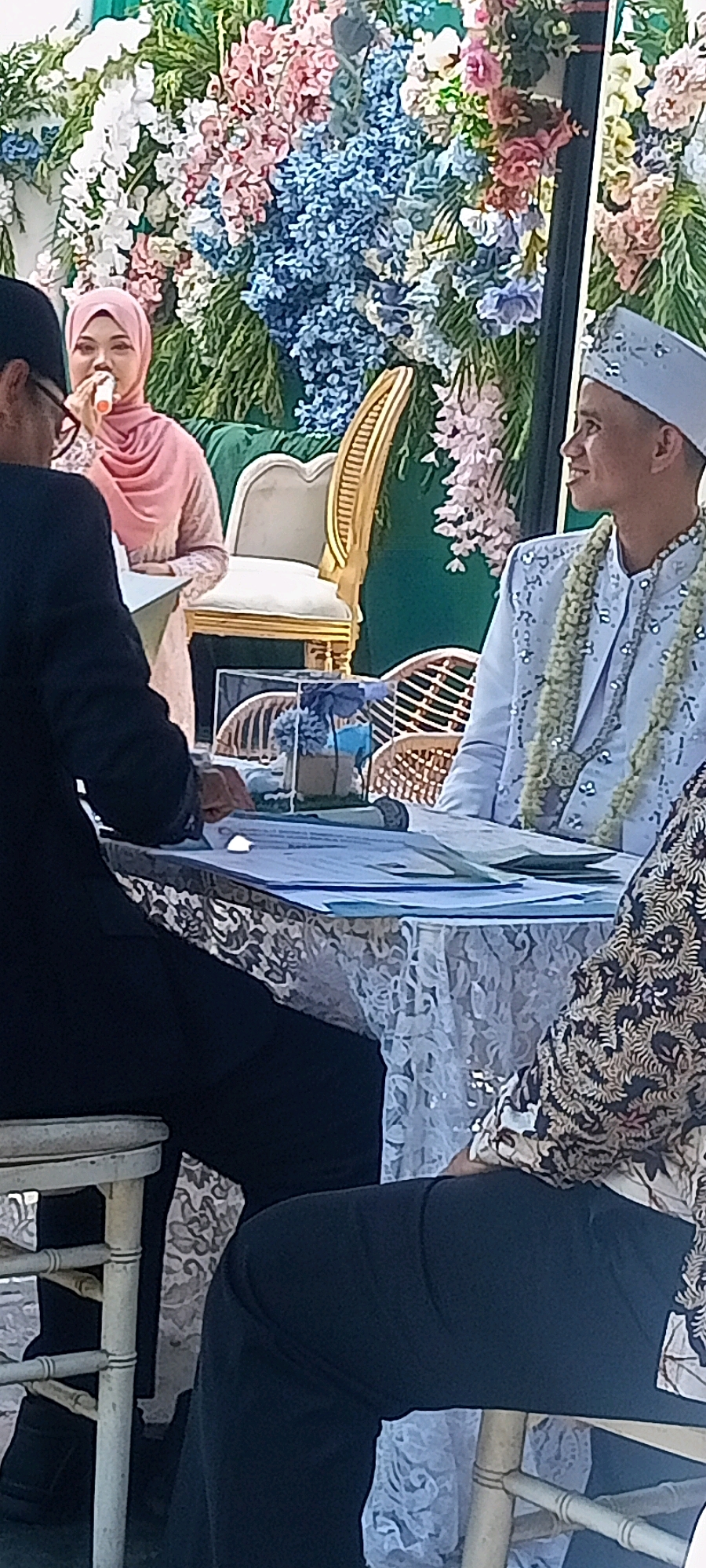 This image depicts a wedding ceremony taking place outdoors, likely in a garden or decorated venue, as indicated by the abundance of flowers in the background. The setup suggests a formal event with a decorated backdrop of white, pink, and blue flowers. In the foreground, a man in a white, embroidered traditional outfit with a matching cap is seated, facing to the right, with a garland of light green flowers around his neck. He appears to be smiling and looking away from the camera. To his left, a woman wearing a pink hijab and a light pink dress is speaking into a microphone, possibly giving a speech or blessing. Another individual in a black suit is partially visible to the far left, seated and looking towards the woman with the microphone. A decorated table with a lace tablecloth is positioned between the seated individuals, holding papers, a microphone, and a small decorative item in a clear container. In the background, an ornate, golden chair with cream upholstery is visible, suggesting a ceremonial seating arrangement. The presence of a batik-patterned garment on a person seated at the table on the right further suggests a cultural event, likely in Indonesia. The specific location within Desa Ponokawan, Indonesia, cannot be definitively determined from the visual information alone, but the overall scene points to a local wedding celebration.