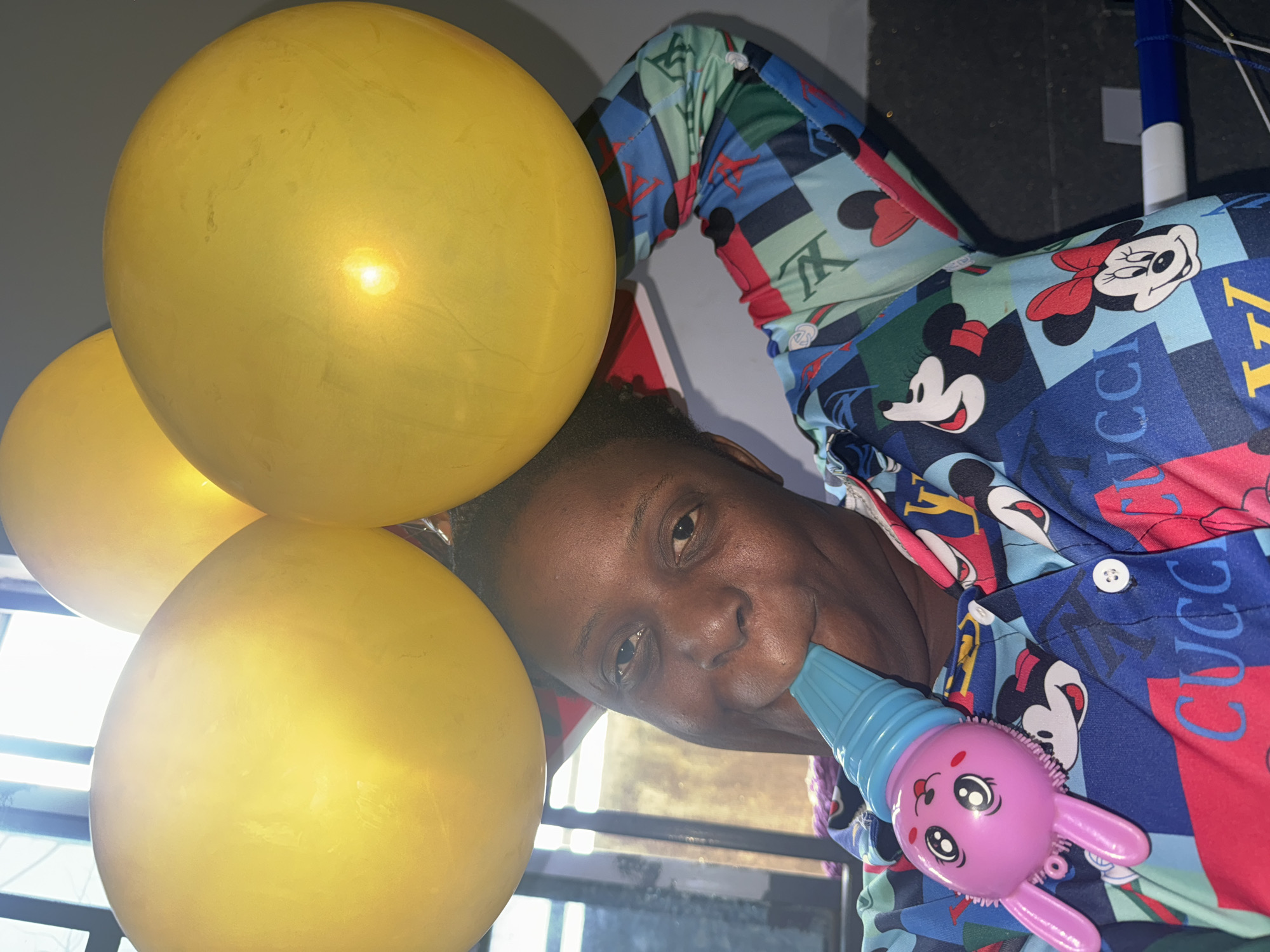 A person with dark hair and dark skin is visible from the shoulders up, holding a pink toy shaped like a bunny in their left hand. They are wearing a colorful shirt with cartoon characters and Gucci logos. Three yellow balloons are positioned to the left of the person's head. The location appears to be indoors, with a window showing sunlight streaming through. The image was probably taken in Sabon Tasha, Nigeria.