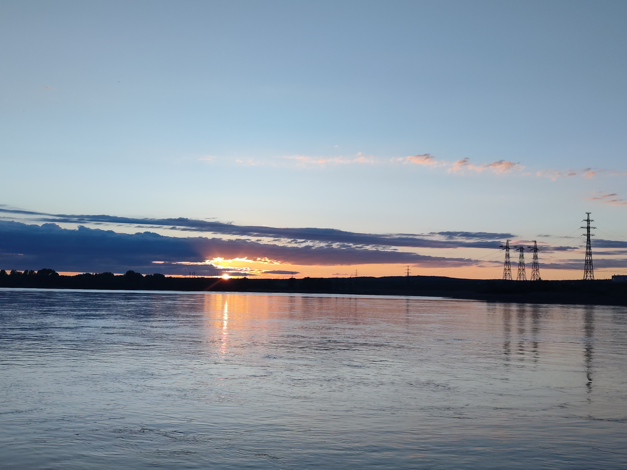 This panoramic image depicts a serene river landscape at sunset or dusk, likely in Chaikovsky, Russia. The sky transitions from a light, clear blue at the top to warm orange, pink, and subtle purple tones near the horizon, illuminated by the setting sun. Dark, heavy clouds stretch across the mid-section of the sky, with the sun's intense orange glow breaking through a gap on the left, casting dramatic light rays upwards and reflecting brightly on the water.
The wide river occupies the foreground and mid-ground, its surface showing gentle ripples and shimmering reflections of the vibrant sky and the sun. The distant bank forms a dark, silhouetted horizon line. A dense line of trees or foliage is visible along this bank. On the right side of the distant bank, several tall metal electricity transmission towers (pylons) stand in stark silhouette against the colorful sky, their distinct shapes mirrored in the water below. The scene is devoid of people or visible text. The overall impression is one of natural beauty and tranquility during the golden hour.