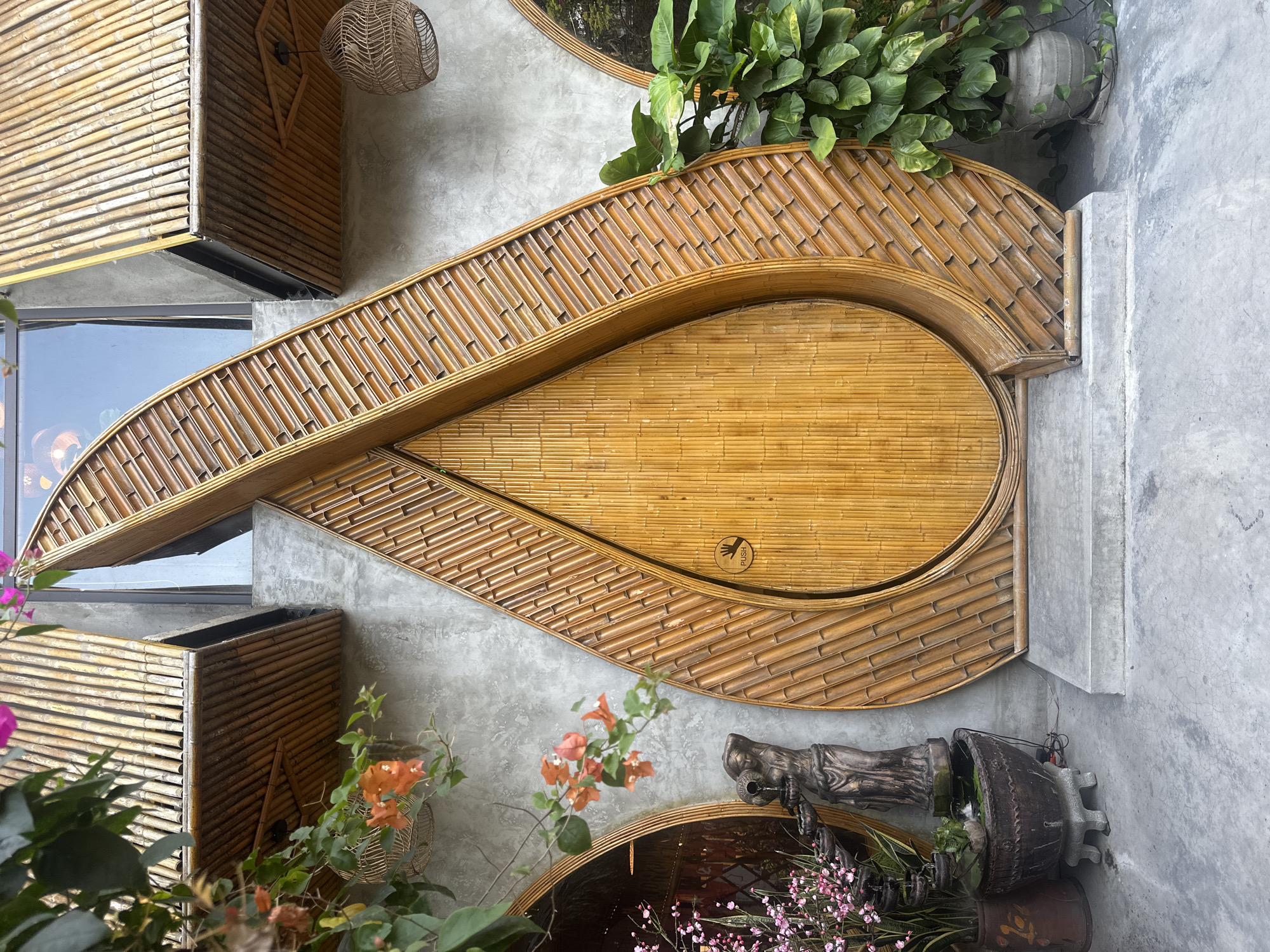 The image depicts an outdoor space in Đà Nẵng, Vietnam, featuring architectural and decorative elements. A large, stylized teardrop-shaped door made of bamboo is the central focus. The door is flanked by a gray concrete wall. Planters with various plants and flowers are positioned near the base of the door, adding greenery to the scene. A window is visible on the left side, while a decorative light fixture hangs near the top. The space appears to be a restaurant or cafe, decorated in a natural style.