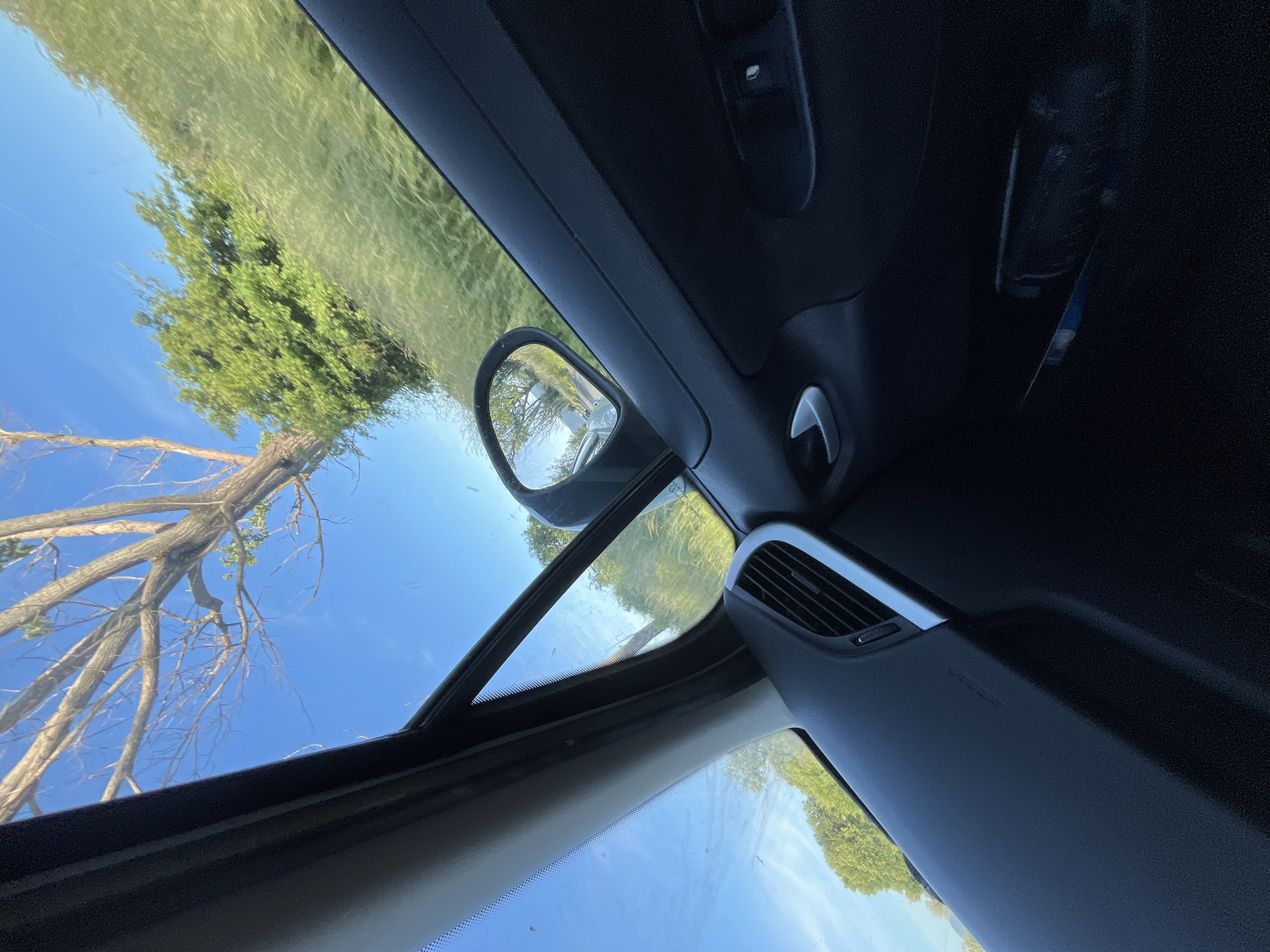 The image shows the interior of a car, looking out through the sunroof. The sky is blue, and there are trees visible. The car's side mirror is also visible. The location is not discernible from the image.