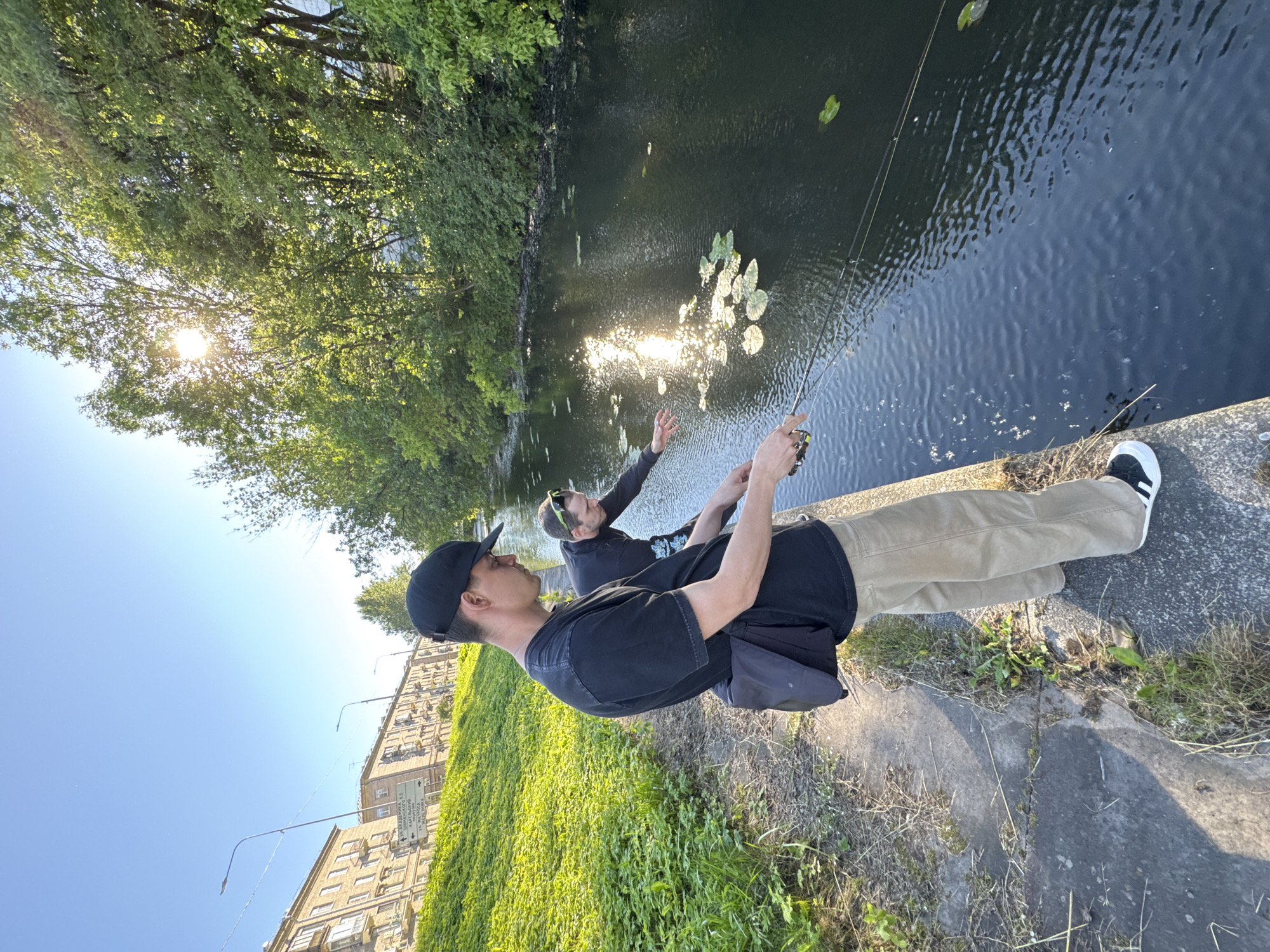 Two young men are fishing on the bank of a canal in Saint Petersburg, Russia. The man in the foreground, wearing a black t-shirt, khaki pants, and a black baseball cap, stands with a fishing rod. He has a dark bag slung over his shoulder. The second man, wearing a black t-shirt and sunglasses on his head, is leaning over the canal and appears to be gesturing towards the water. Lily pads dot the dark water, and the sun reflects brightly on its surface. Behind the men, a grassy embankment slopes up towards a street lined with apartment buildings and trees. The sky is clear and blue, indicating a sunny day.