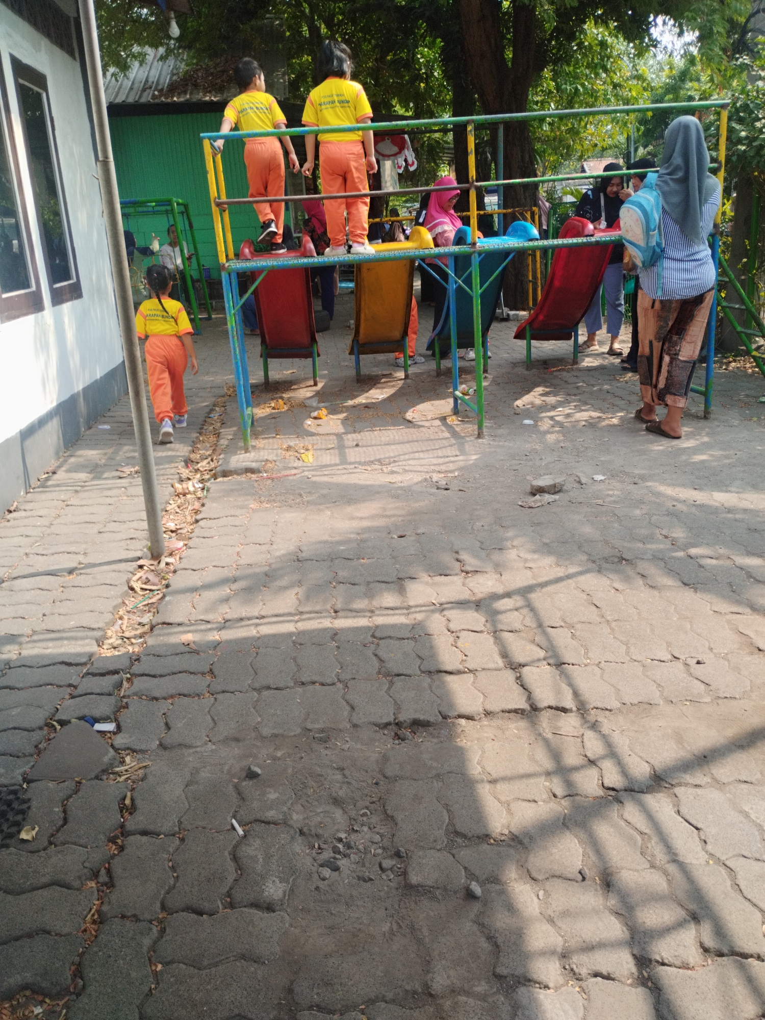 The image depicts an outdoor playground area, likely part of a school or community center, under bright daylight. The ground is paved with interlocking grey concrete blocks, some of which are cracked and uneven, with debris and dry leaves scattered in the crevices. To the left, a white building with dark-framed windows is visible.
In the center of the frame is a colorful playground structure with red and yellow slides, blue and green metal supports, and seats. Several individuals are present. Two children, wearing matching yellow t-shirts with text and orange pants, are standing on the elevated platform of the playground. Another child, also in a yellow t-shirt and orange pants, is walking away from the playground structure towards the foreground.
Several adults are also present. One adult, wearing a grey hijab and a blue and white striped shirt with patterned pants, is facing away from the camera, carrying a light blue backpack. Another adult, wearing a dark hijab, is standing near the red slide. Two other individuals, one in a pink hijab and another in a black top, are also near the playground. The overall impression is one of a busy playground scene with children and adults interacting or observing.
The environment is characterized by lush green trees providing shade in the background. The lighting suggests it is mid-day, with distinct shadows cast on the paved ground. The presence of outdoor recreational equipment and the attire of the people suggest a warm climate. Given the context of the bright sun and vegetation, this scene is consistent with an outdoor location in a city like Surabaya, Indonesia, which experiences a tropical climate.