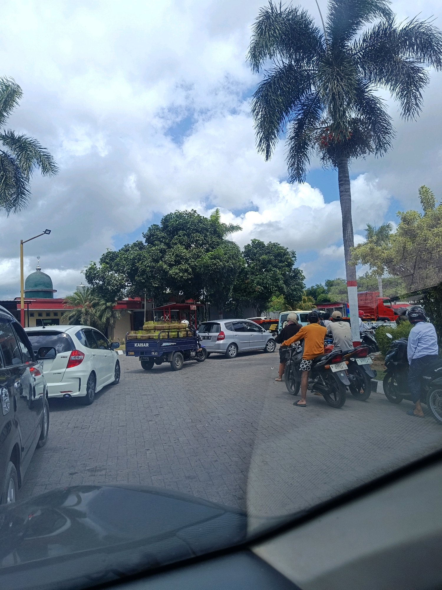 This image shows a street scene in Tempeh Lor, Indonesia, likely during daylight hours given the bright sky and visible shadows. The foreground is dominated by the hood and windshield of a vehicle, suggesting the photo was taken from inside a car. The road is paved with interlocking bricks.
In the mid-ground, a white Honda Jazz is parked on the left, facing away from the camera. To its right, a blue three-wheeled utility vehicle, branded "KAISAR," is carrying a load of what appear to be green logs or stalks. Further down the street, several motorcycles are lined up, some with riders. One motorcycle has a rider wearing an orange shirt and patterned shorts, another has a rider in a white shirt. A person in a light blue shirt is seated on a motorcycle on the far right.
In the background, a mosque with a green dome and red accents is visible. Lush green trees and palm trees are abundant throughout the scene, with a particularly tall palm tree on the right side of the frame. The sky is partly cloudy with patches of blue. A red truck is parked behind some vegetation on the right. The overall impression is a busy, tropical urban environment.