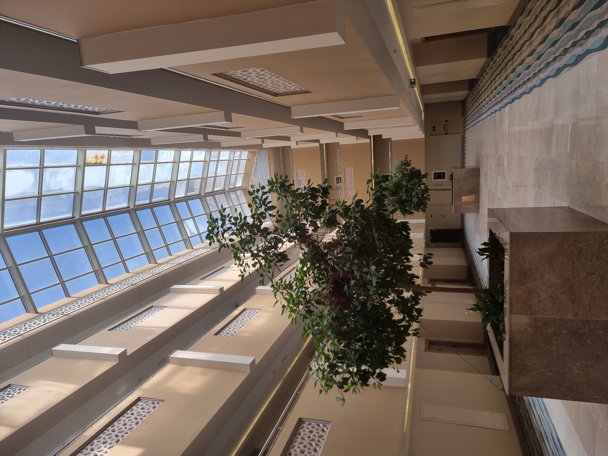 An indoor atrium with a large glass ceiling features several stories of balconies overlooking a central open space. The walls are painted a light tan color, with white architectural accents and decorative patterned panels. Two potted trees are placed in the open area, with a larger tree in the foreground. The floor is tiled with a light marble pattern and features a patterned rug runner along one side. Doors are visible on the balconies and at the end of the corridor. The scene appears to be a modern hotel or office building.