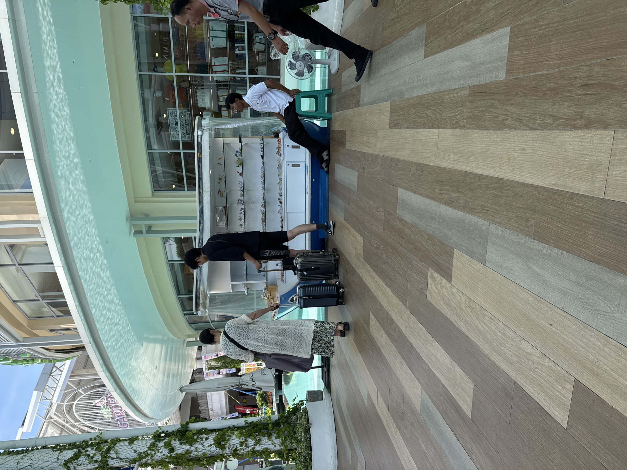 In Patong, Thailand, a scene unfolds outside a retail establishment with a curved canopy. Three men and one woman are present. A woman in a long, white patterned dress with a floral skirt walks away from the viewer, pulling a dark suitcase on wheels. Beside her, a man in black shorts and a t-shirt pulls another dark suitcase. A second man, wearing a white polo shirt and black pants, sits on a green stool. To the right, a man in black pants and a grey t-shirt walks towards the viewer. In front of the retail establishment, a display case filled with small items is visible. A white fan sits on the ground to the right of the man on the stool. The ground is a wooden-planked walkway. In the background, the building features large windows and signage.