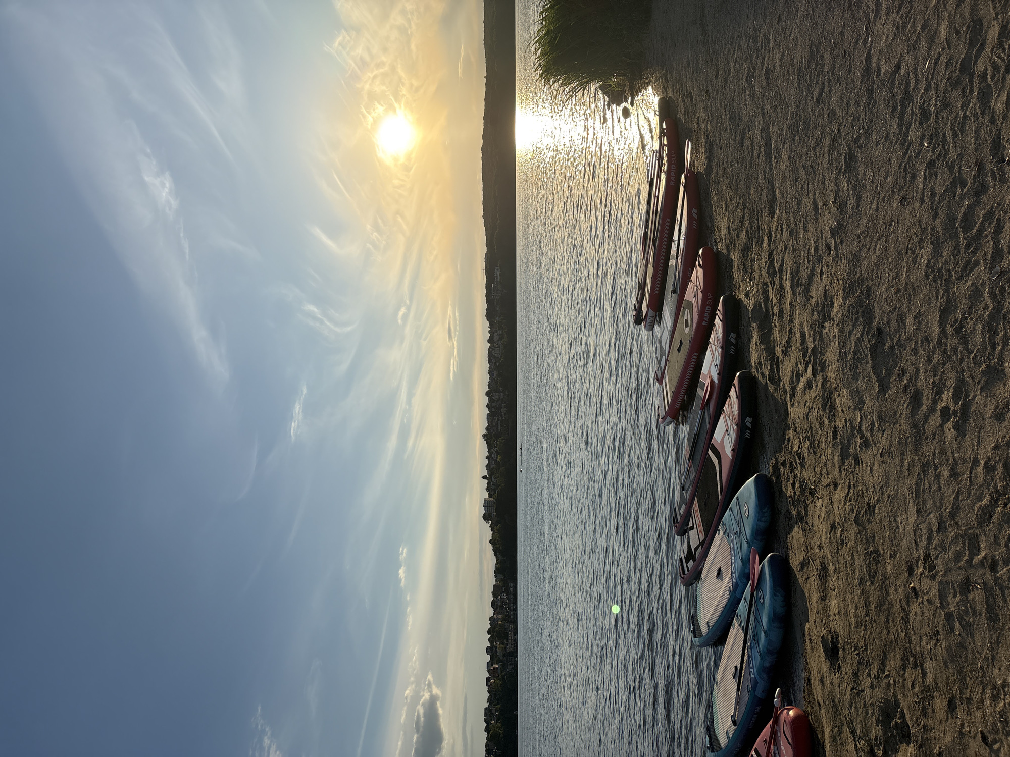 The image depicts a serene sunset scene at the Ternopil lake in Ternopil, Ukraine. Several paddleboards are neatly arranged on the sandy shore, facing the tranquil waters. The setting sun casts a warm, golden glow across the lake, creating a shimmering reflection. The sky is adorned with wispy clouds, adding to the picturesque ambiance. In the distance, the city skyline is visible, with buildings and lush greenery lining the horizon. A few people can be seen in the water, possibly enjoying the last moments of daylight. The overall atmosphere is peaceful and inviting, showcasing the natural beauty of Ternopil.