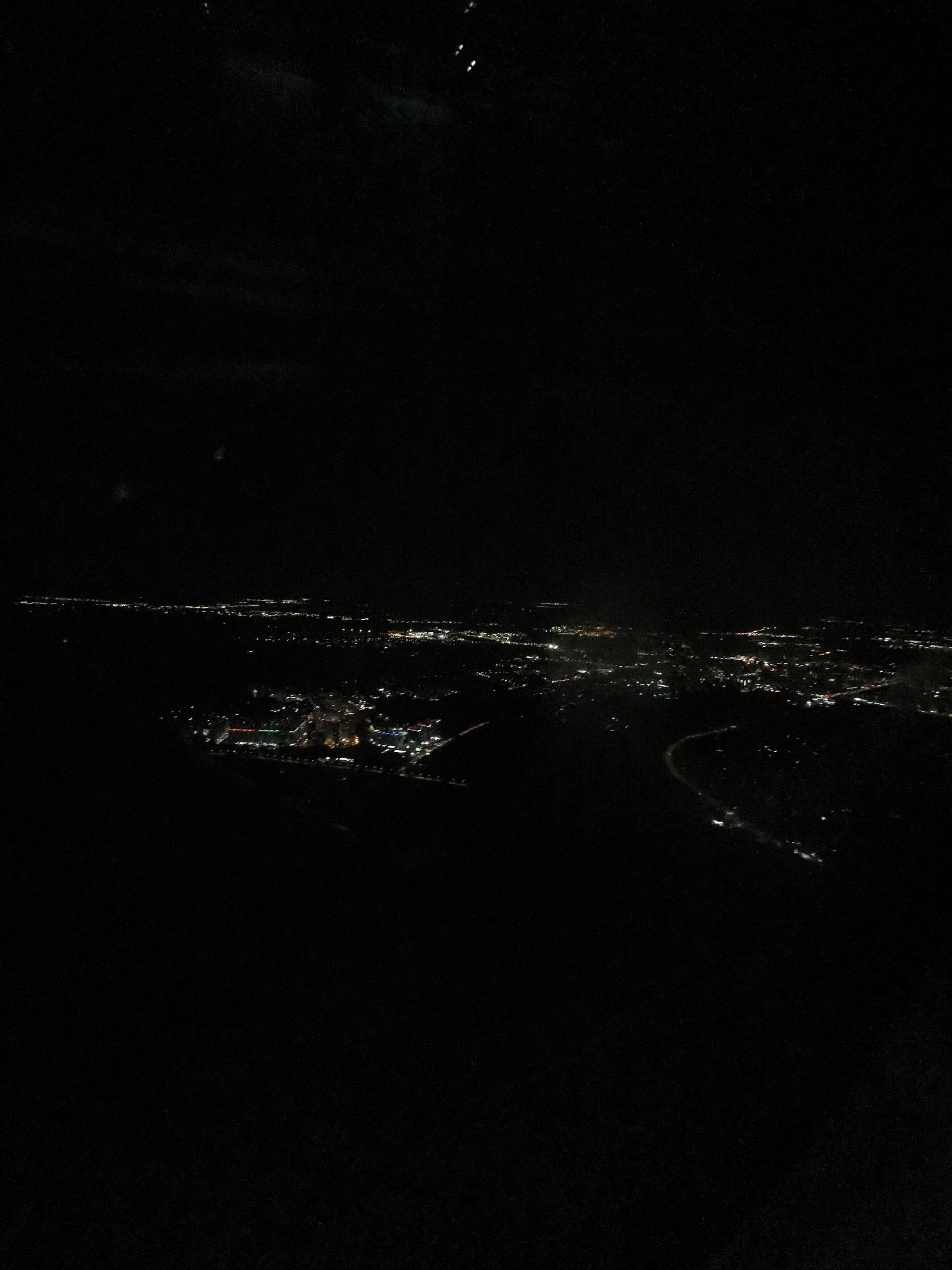 The image depicts a nighttime view of a city from an elevated perspective, possibly from an airplane. The landscape is primarily dark, with clusters and lines of lights indicating urban areas and roadways. In the lower left portion of the frame, a concentrated area of colored lights suggests a distinct complex or facility. A curving road is visible on the right side. The specific location is Khimki, Russia.