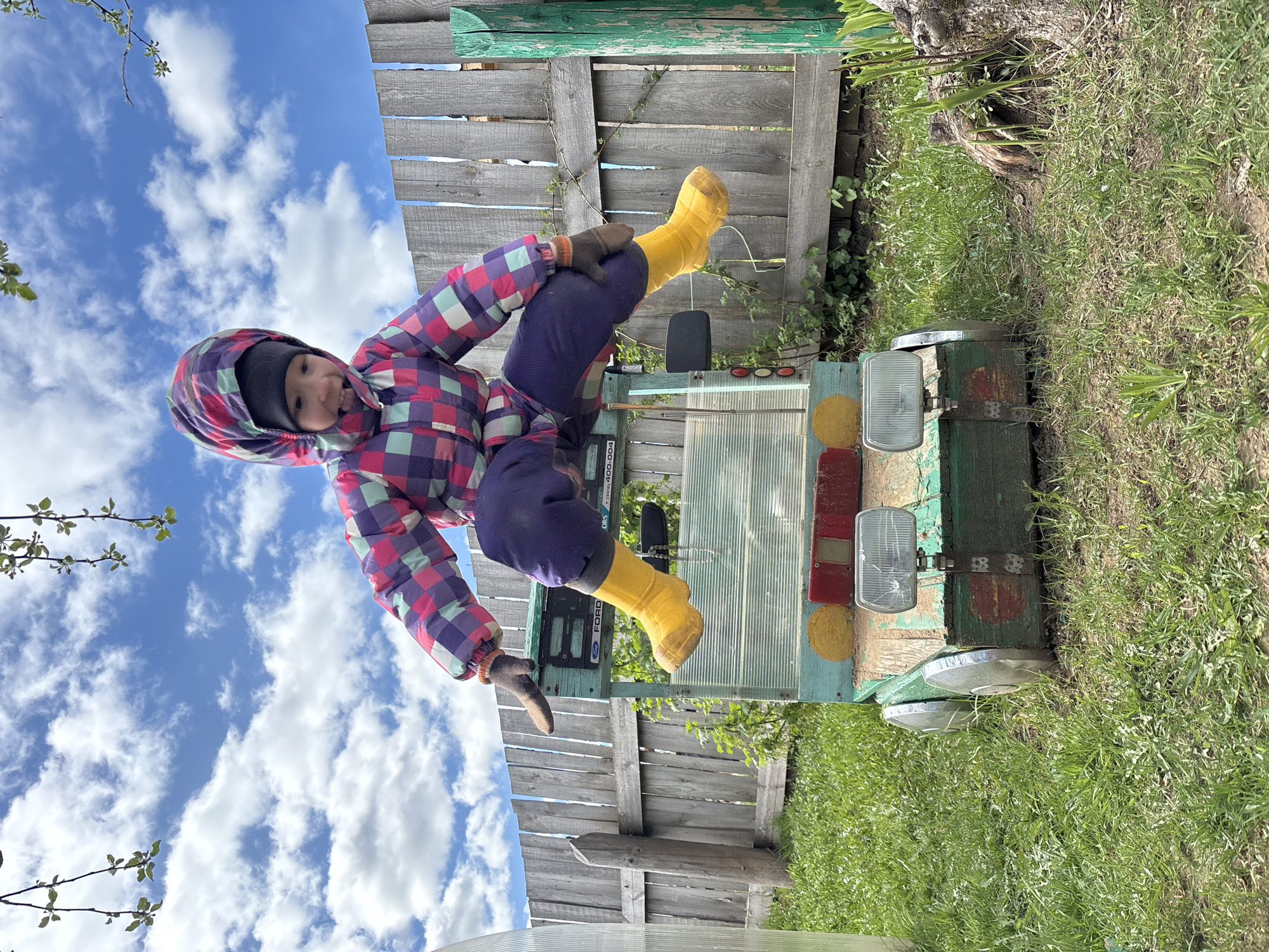 A child wearing a multi-colored checkered jacket, navy pants, and yellow boots is sitting on a green and silver vehicle with headlights, presumably a tractor or similar agricultural machine. The background includes a wooden fence, a sky with white clouds, and a grassy area. The environment suggests a rural or suburban setting. A sign on the vehicle reads "FORD 4000." The location is in Ваньки, Russia.