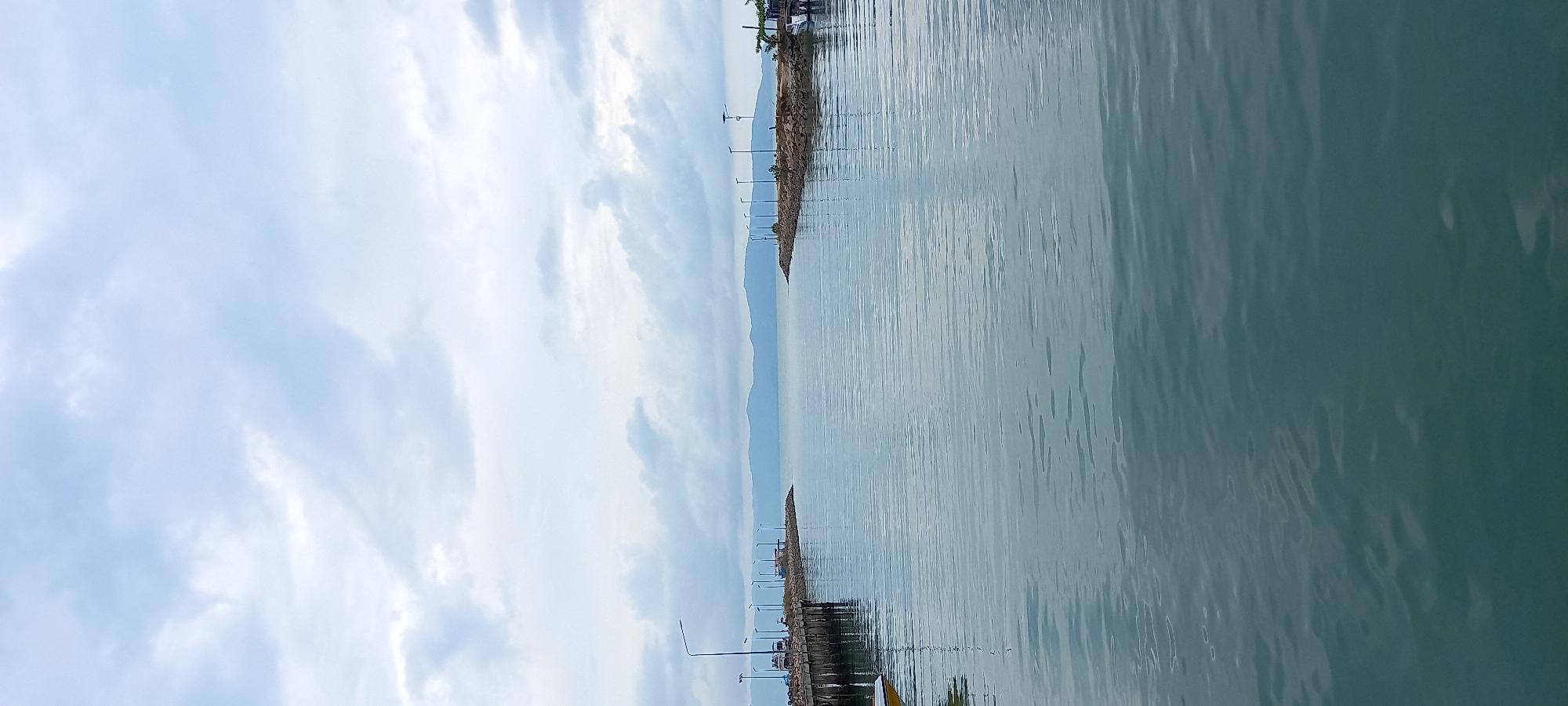 The image captures a seaside view in Baan Tai, Thailand. The scene is dominated by water, its surface displaying subtle ripples and a muted teal color. A rock breakwater stretches across the horizon, with spaced light poles lining its edge. Behind the breakwater, a strip of land is visible, with distant mountain silhouettes suggesting a mainland presence. Above, the sky is filled with an array of white clouds against a pale blue backdrop. The image is oriented in portrait style.