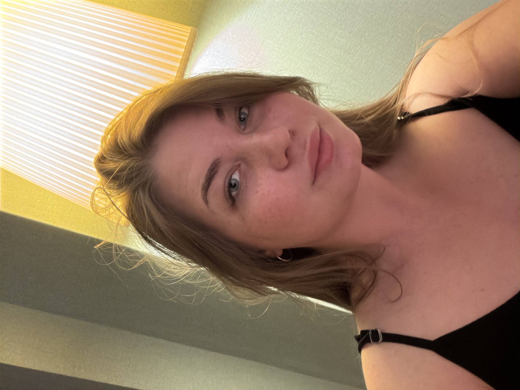 A forward-facing selfie-style photograph captures a young adult female, approximately in her late teens or early twenties, positioned indoors within Kazan, Russia. The subject, with fair skin, blue eyes, and light brown hair, looks directly at the camera with a soft, closed-lip expression. Freckles are visible across her cheeks. She is wearing a black top with thin straps, one of which displays a silver-toned metal adjuster on her left shoulder. A small, simple silver hoop earring is present on her left earlobe. Her hair is illuminated by a warm, bright light source originating from the upper right, suggesting artificial lighting. The background consists of interior walls. To the upper right, a prominent, vertically ribbed lampshade emits a strong, warm yellow light, illuminating a light green-yellow wall section. To the upper left, the wall features subtle vertical striations or panels in shades of grey and beige.