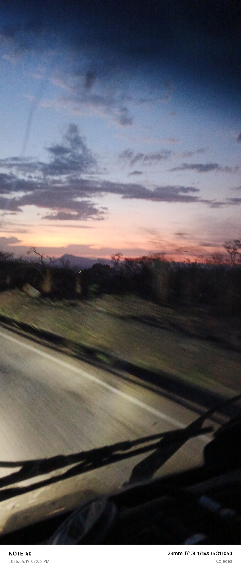 The image depicts a view from inside a vehicle, likely a car, while in motion on a road during a sunset. In the foreground, the windshield wipers are visible. The road, with a white line marking the edge, stretches into the middle ground. Beyond the road, a blurred landscape with trees and vegetation suggests a rural or suburban setting. The sky is dominated by a colorful sunset, transitioning from blue at the top to orange and pink near the horizon. The image seems to be taken in motion, as evidenced by the motion blur. The image was taken with a 23mm f/1.8 lens, 1/14s exposure, ISO 11050. The location provided, La Galera, Venezuela, suggests this is where the image was taken. There are no identifiable people.