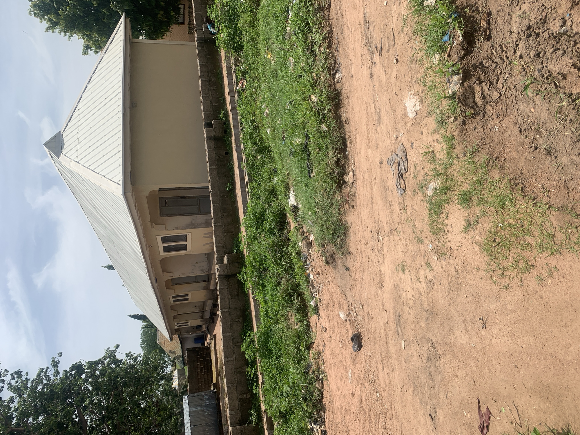 The image depicts a single-story building with a light-colored exterior and a corrugated metal roof, a common architectural style in Jauro Yinu, Nigeria. The building appears to be a residential or commercial establishment, with several doors and windows visible along its facade. In front of the building, a low wall made of concrete blocks separates it from a dusty path and an area with overgrown green vegetation. The ground is predominantly dirt, with scattered debris, including a single black shoe, some fabric, and plastic waste, indicating a somewhat unkempt environment. The sky above is partly cloudy, suggesting fair weather conditions. The scene is devoid of any visible people or animals, contributing to a quiet and still atmosphere. The overall impression is of a typical street scene in a Nigerian town, characterized by simple structures and an outdoor environment that reflects the local climate and living conditions.
