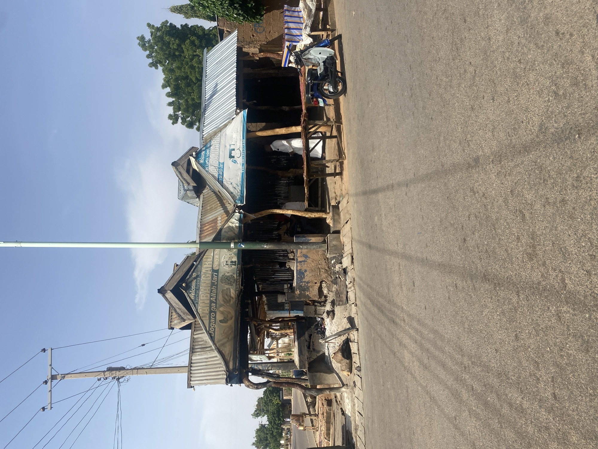 The image depicts a roadside scene in Gombe, Nigeria. A corrugated metal structure serves as a shop or stall, indicated by a sign reading "DANBANGARU" and the words "Jogaro ga Allah Jari Suya Si," suggesting it sells suya (grilled meat). The structure appears weathered and possibly damaged. A motorcycle is parked near the shop. A utility pole with electrical wires is visible, casting shadows on the paved road. The sky is clear, indicating daytime. The environment seems dusty and urban.