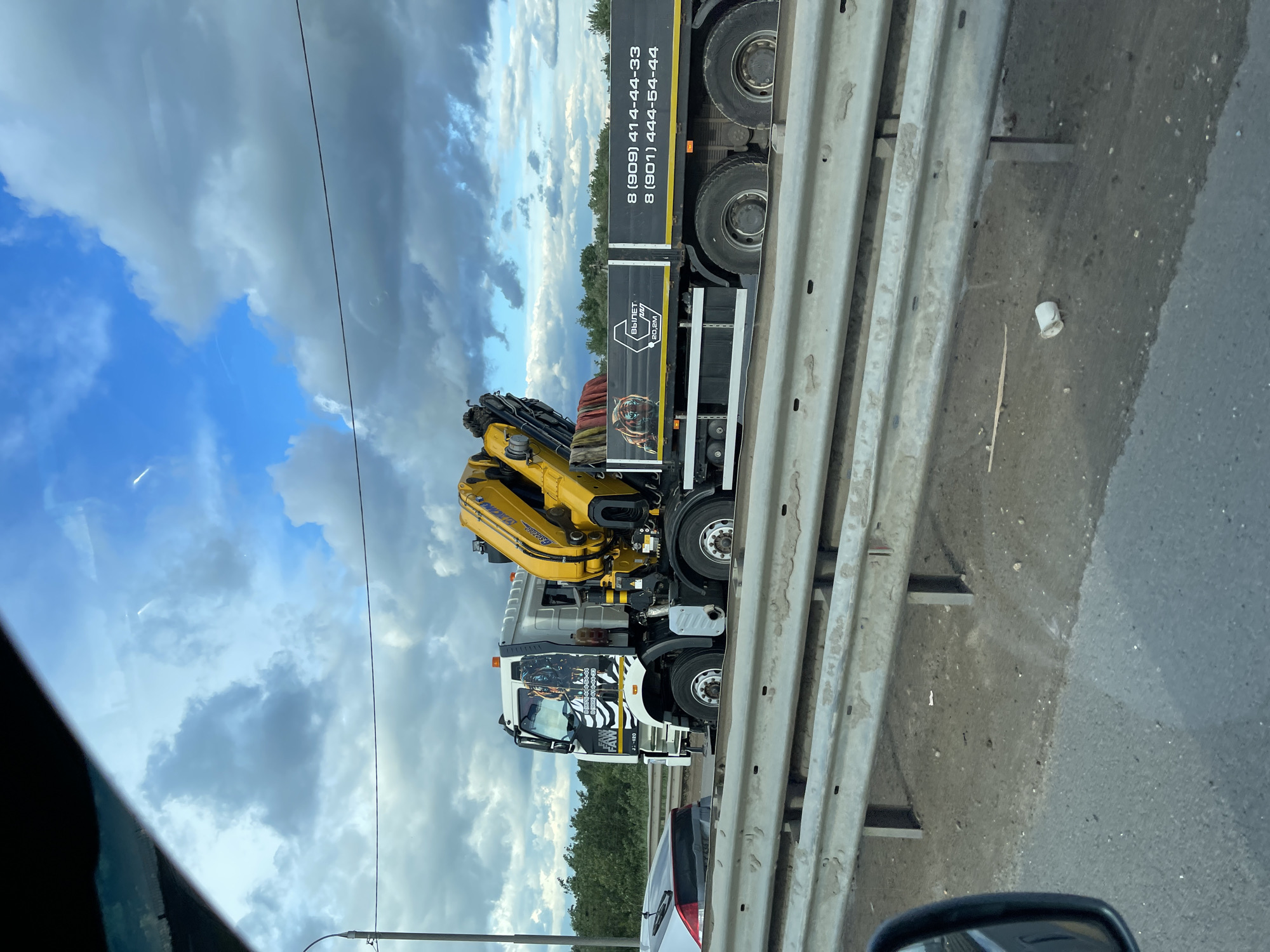 The image shows a truck-mounted crane stopped on the side of a highway in Unknown, Russia. The crane is yellow and mounted on a white truck. The truck has a black banner with Russian phone numbers: 8 (909) 414-44-33 and 8 (901) 444-54-44. A guardrail separates the truck from the traffic lanes. The sky is cloudy, with patches of blue. The image was likely taken from inside a car. Other cars are visible near the truck. A white cup is visible near the guardrail.